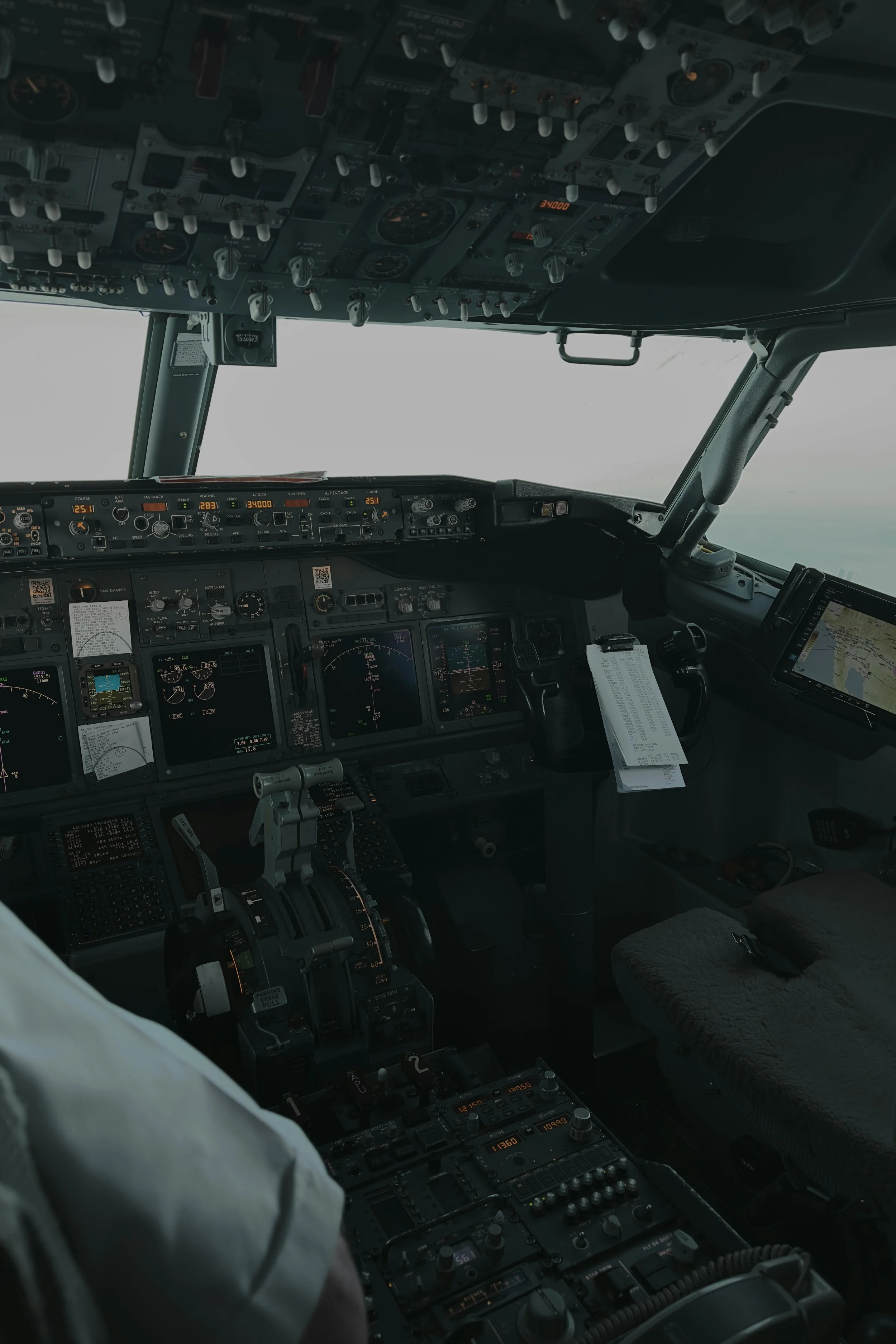 Cockpit of an airplane with various controls, screens, and instruments, with overcast sky visible through the windshield.