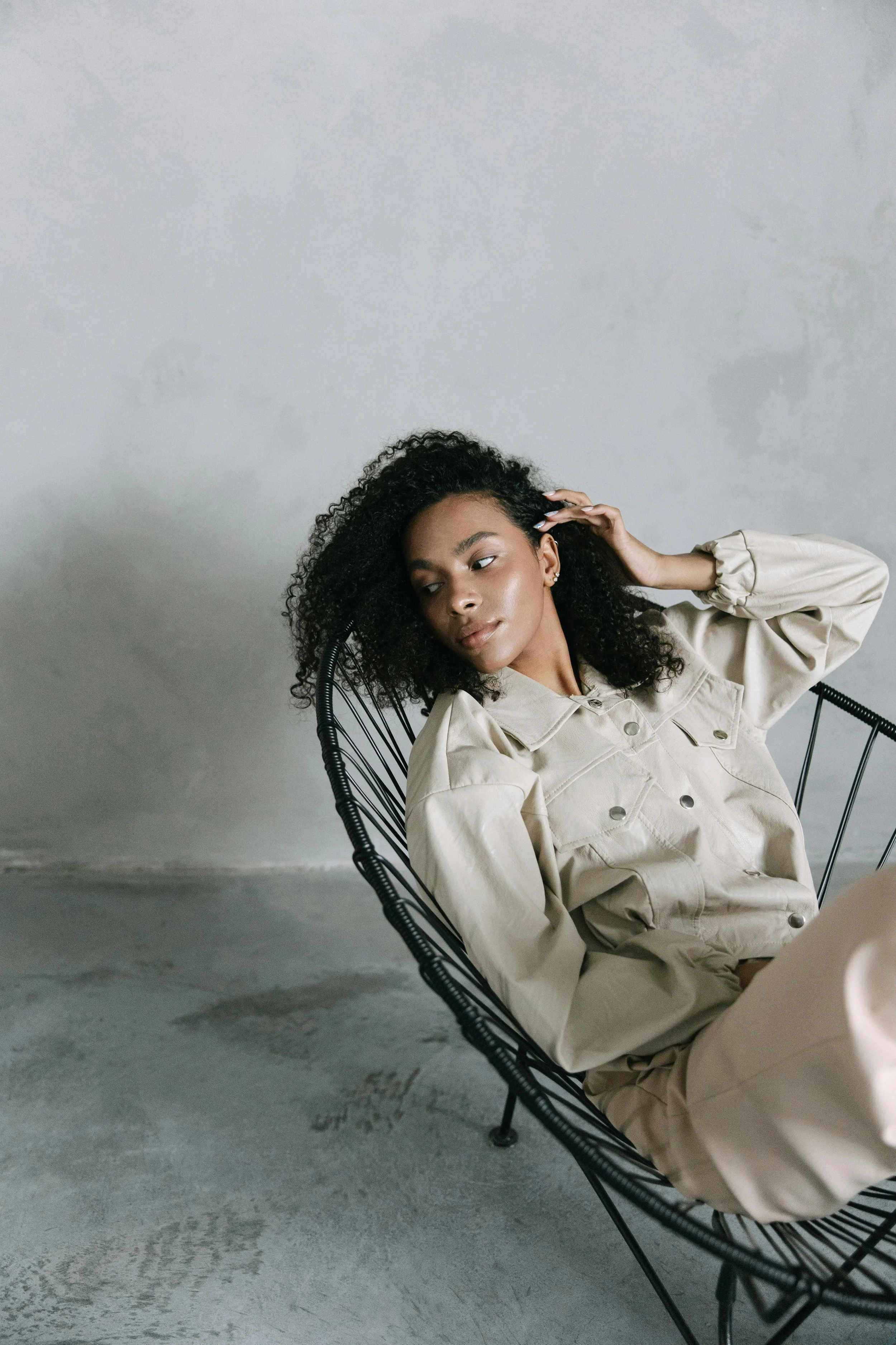 A woman with curly black hair, wearing a beige jacket and matching pants, sitting in a black wire chair against a light gray textured wall, looking to the side with a relaxed expression.