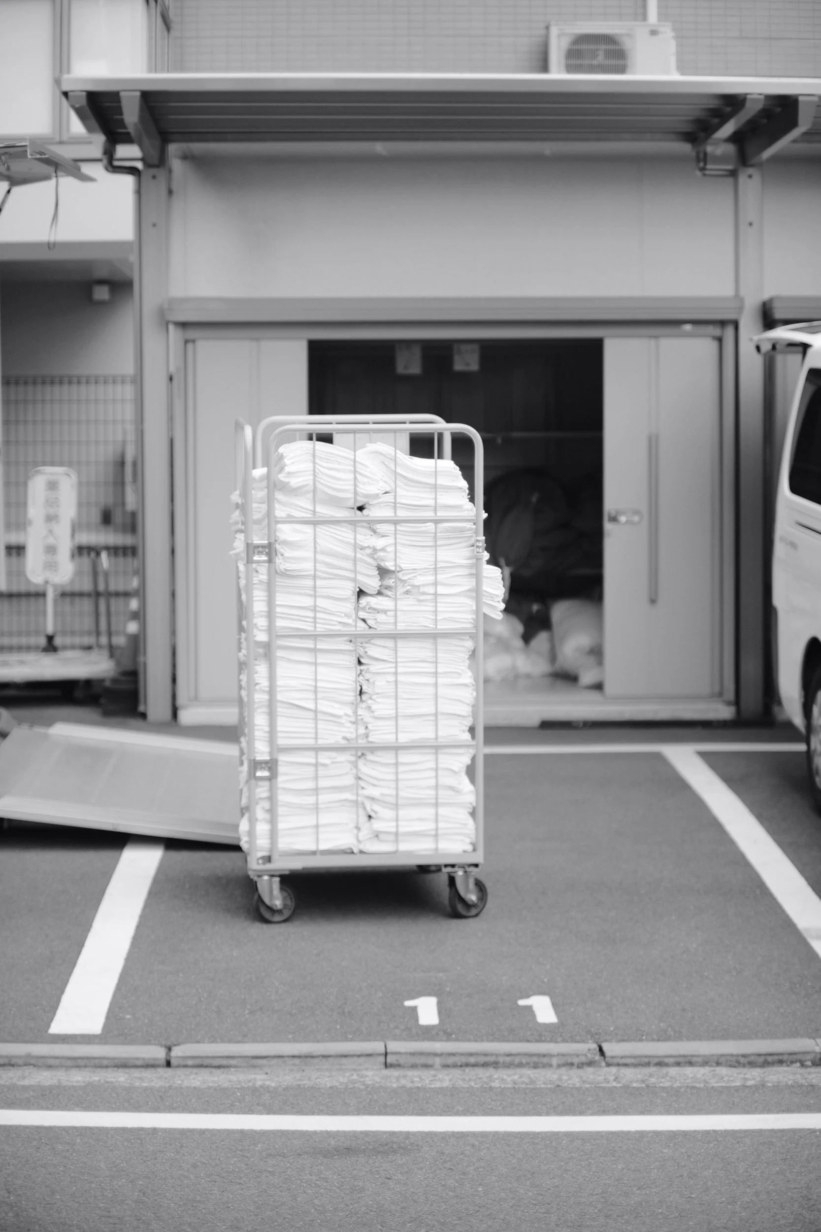 Ein zerlegbarer Wäschwagen voller weißer Handtücher oder Kleidung, vor einer Lager- oder Lieferhalle, im Parkhaus. Das Bild ist schwarz-weiß.