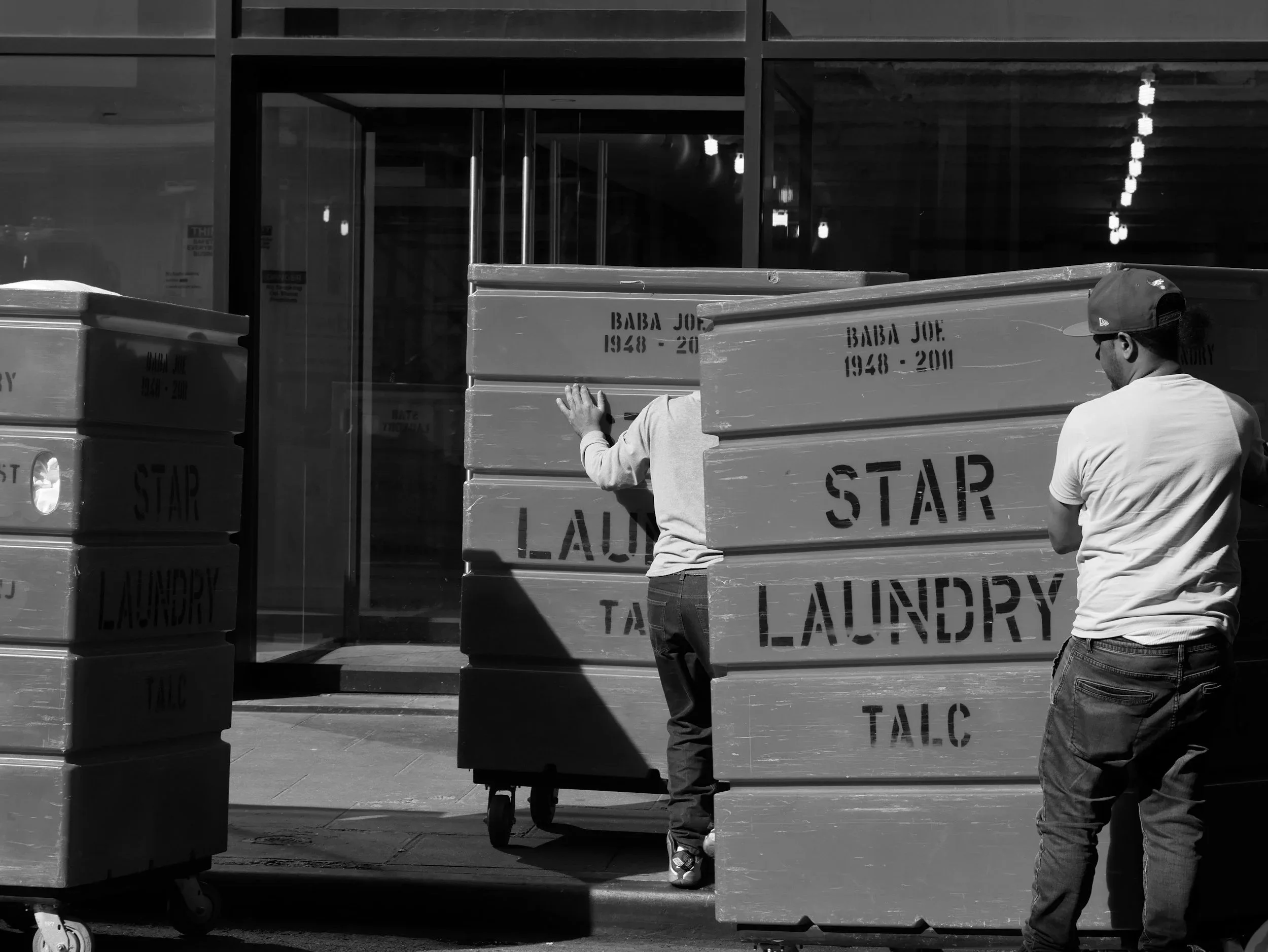 Zwei Menschen setzen große Kisten mit der Aufschrift "Star Laundry" im Stadtbild ab, eine Person mit Baseballkappe beobachtet, während eine Person mit kurzen Haaren die Kiste schiebt.