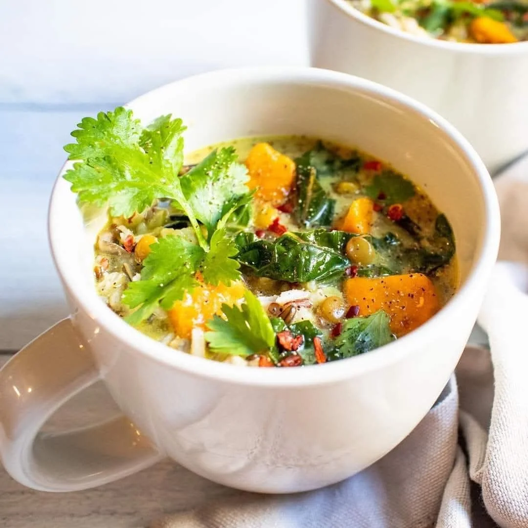 A white mug filled with vegetable soup garnished with cilantro, containing chunks of sweet potato, spinach, and herbs. In the background, there is another bowl of soup.
