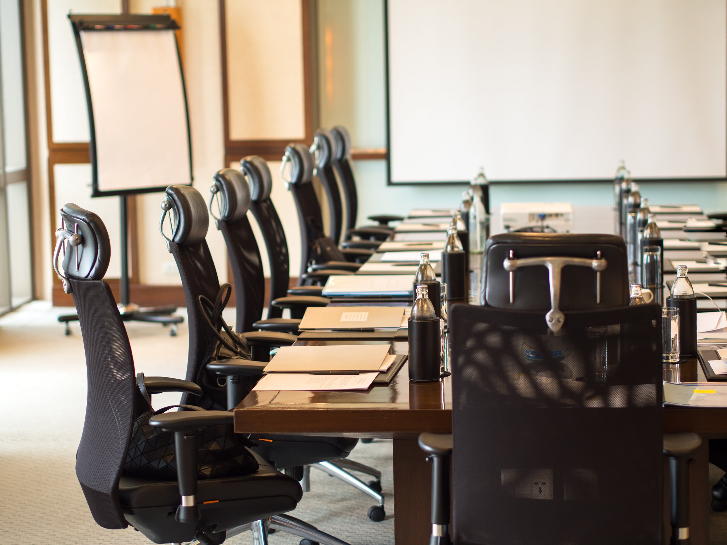 Empty conference room with black office chairs around a long wooden table, with bottles of water, notebooks, and papers, in front of whiteboards and projection screens.
