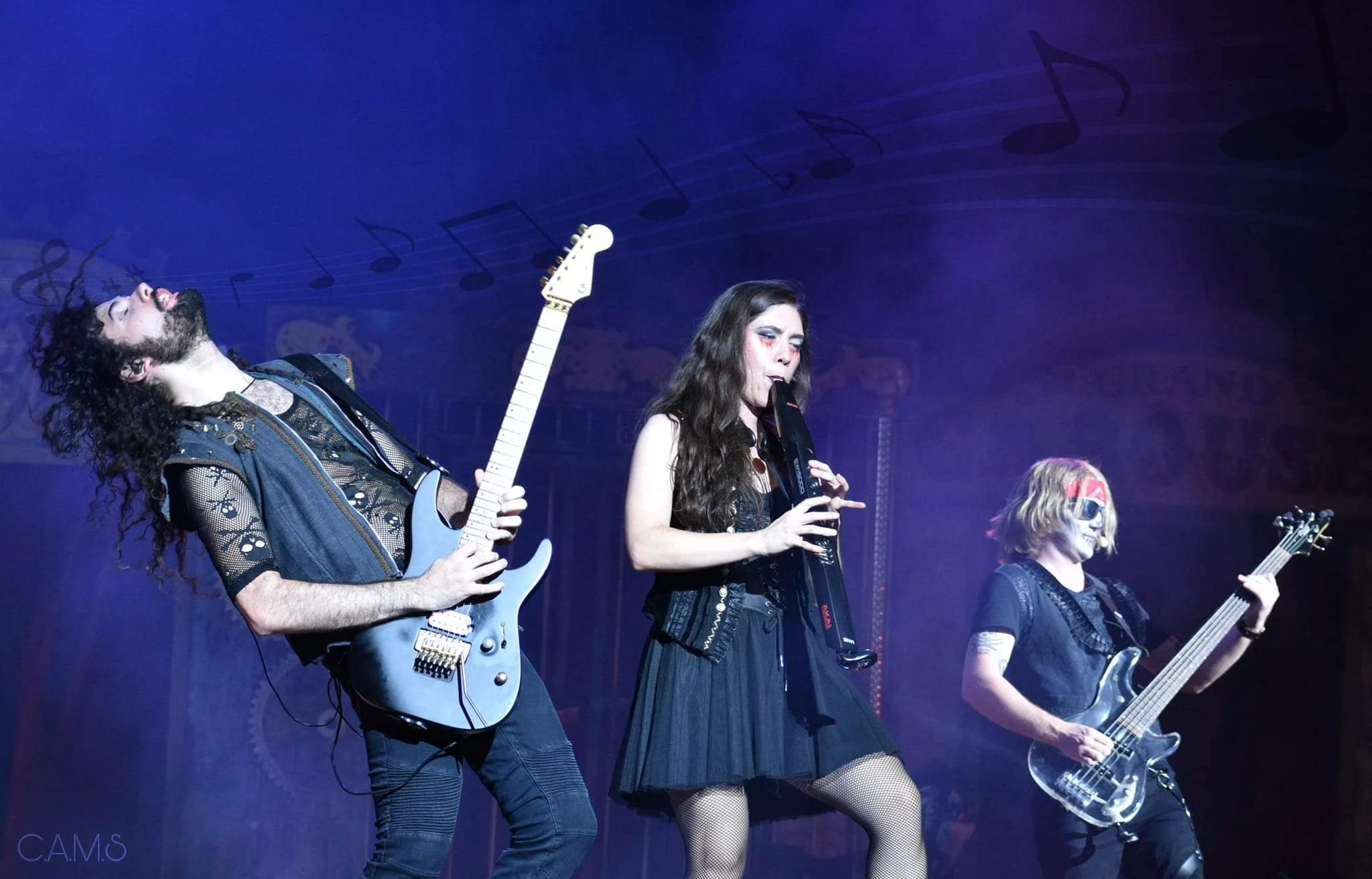 Three performers in black gothic rocker costumes play guitar and sing under blue stage lighting for Dr. Diabolical's Monster Mash Bash