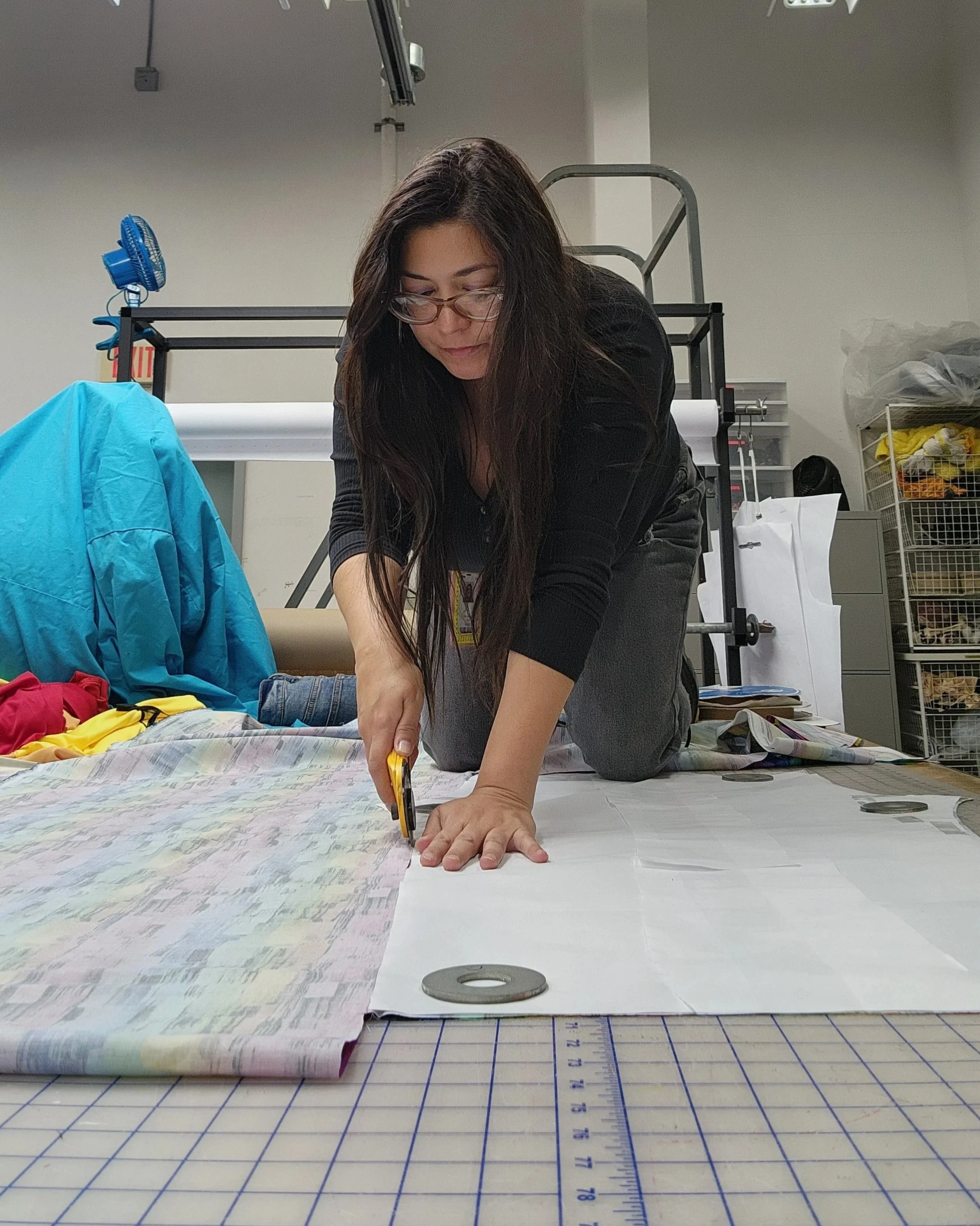 A woman with long dark hair and glasses is kneeling on the floor, cutting fabric on a large cutting mat, surrounded by fabric, tools, and measuring equipment in a workshop or studio.