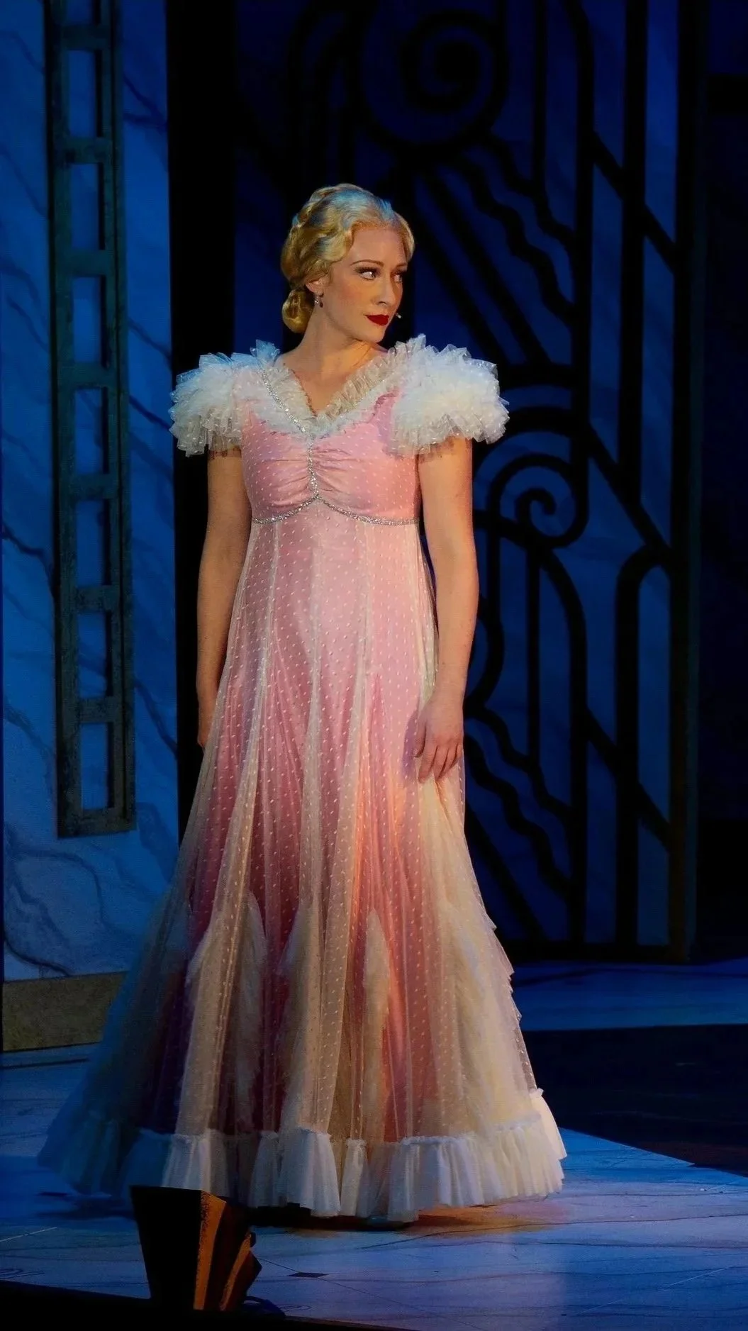 A woman in a long pink dress with ruffled shoulders and polka dots standing on a stage with a dark background.
