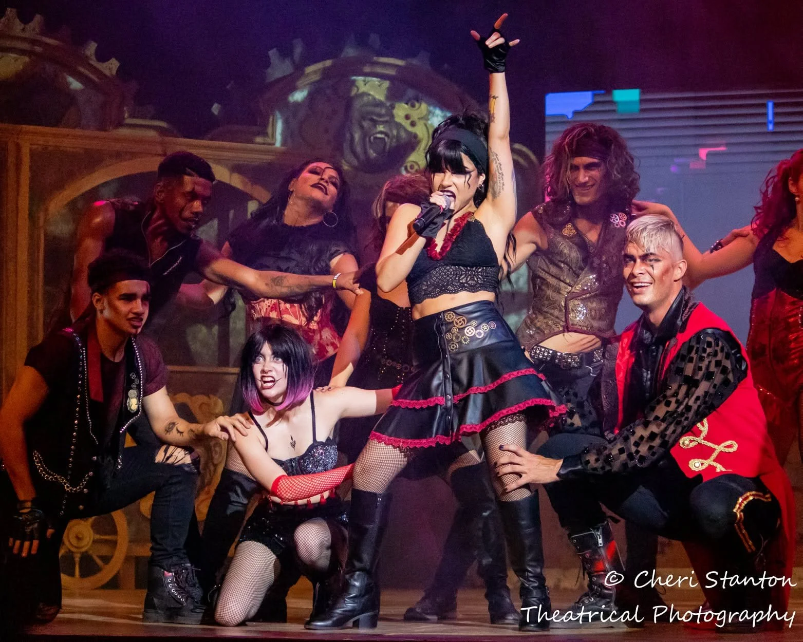A group of performers in red and black gothic costumes celebrate together on an ornate steampunk-style set for Dr. Diabolical's Monster Mash Bash