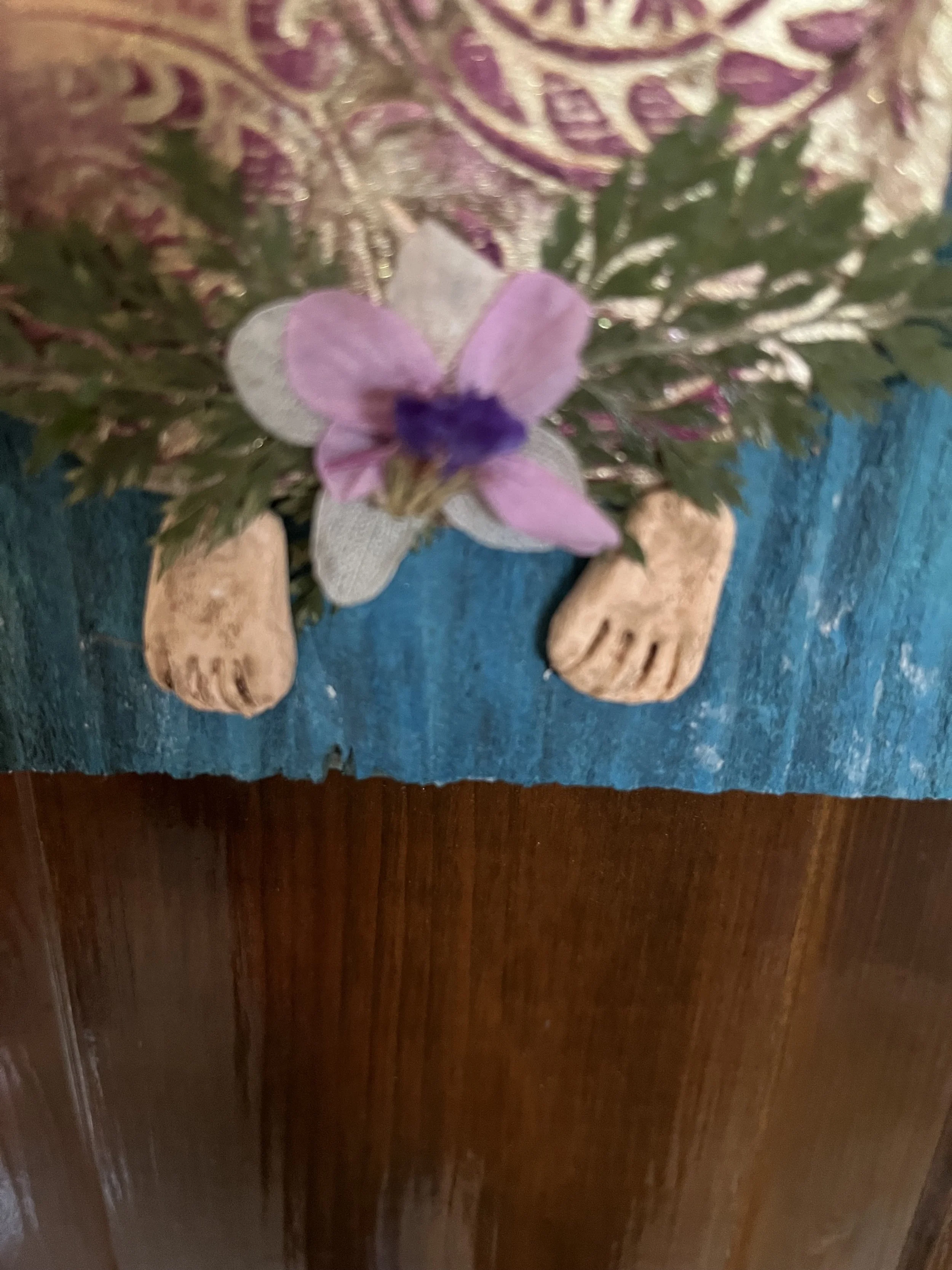 Decorative art piece with small feet, purple flowers, green leaves, on a textured background.