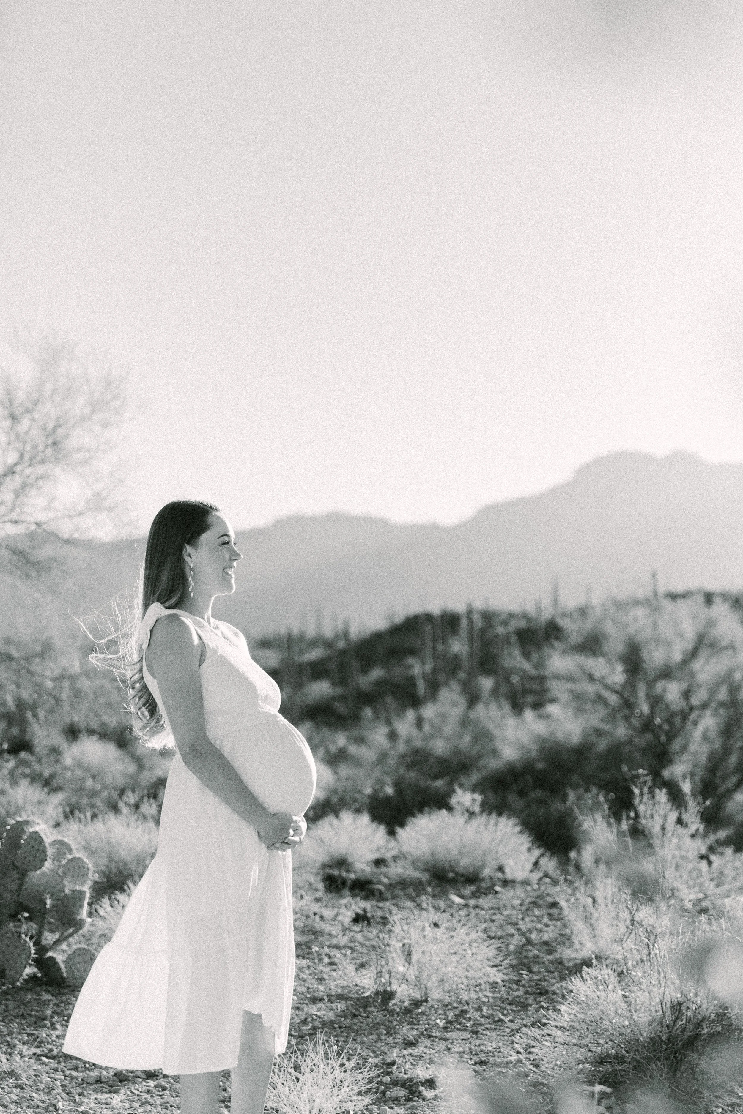 A pregnant woman smiling and holding her belly while standing outdoors in a desert landscape with mountains in the background, in black and white.