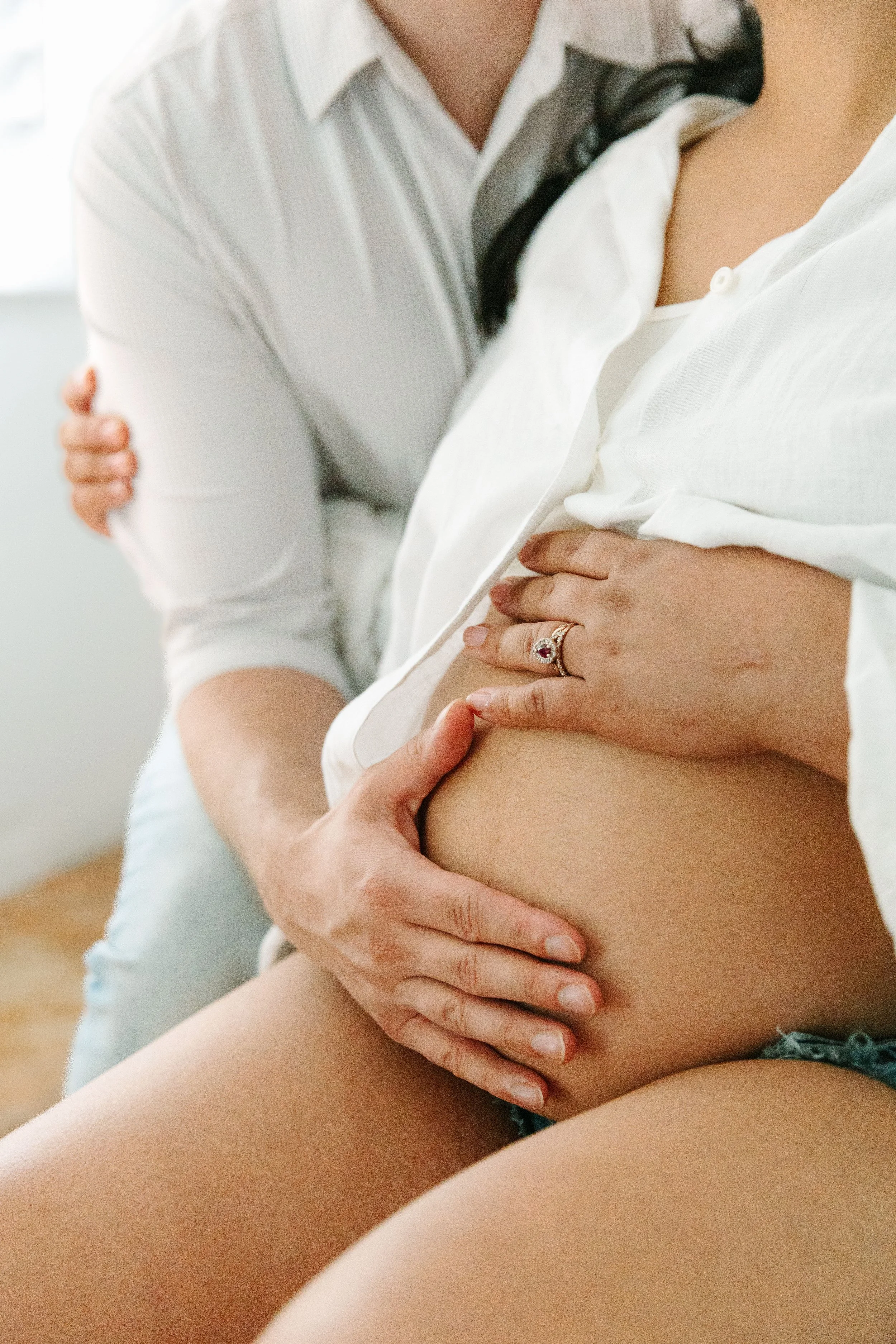 A close-up of a woman holding her pregnant belly while sitting, with a man behind her gently touching her stomach. The woman is wearing a white shirt, and the man is wearing a striped shirt.