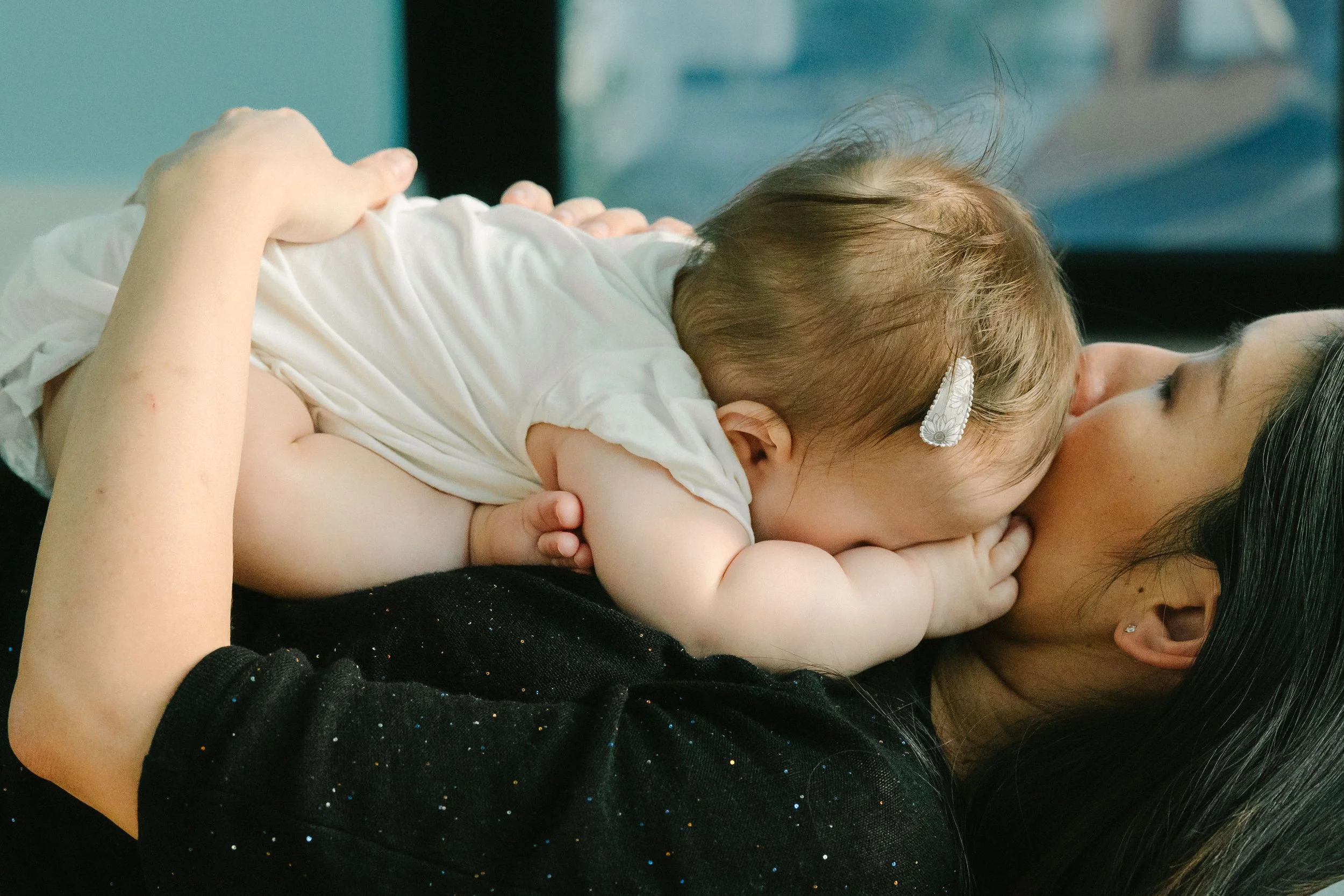 A baby lying on a woman's chest, breastfeeding, with the woman lying on her back. The baby is wearing a white outfit and has a hair clip. The woman has long dark hair and wears a black top with small colorful specks.