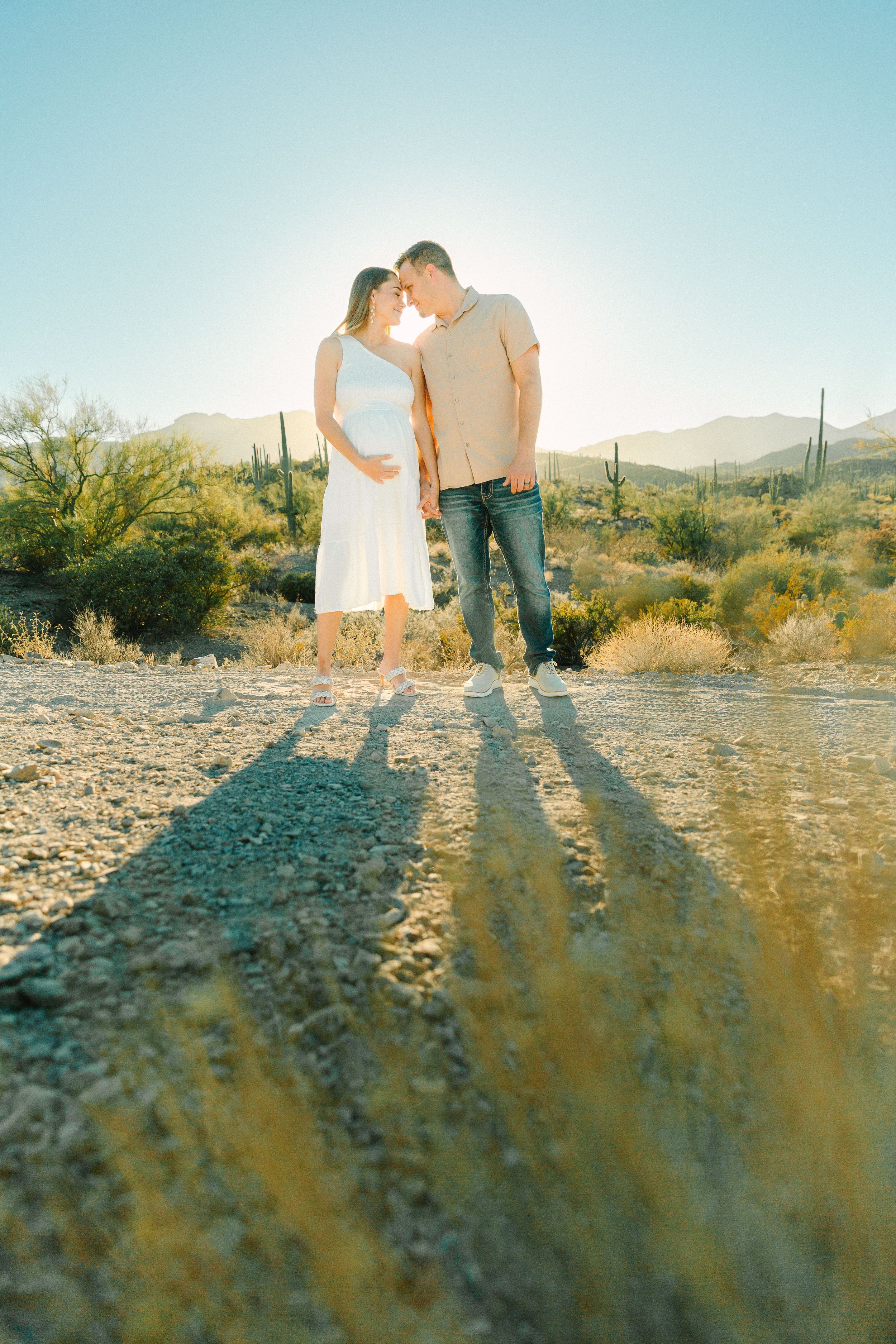 A couple standing close together with their foreheads touching, holding hands in a desert landscape with cacti and mountains in the background during sunset.