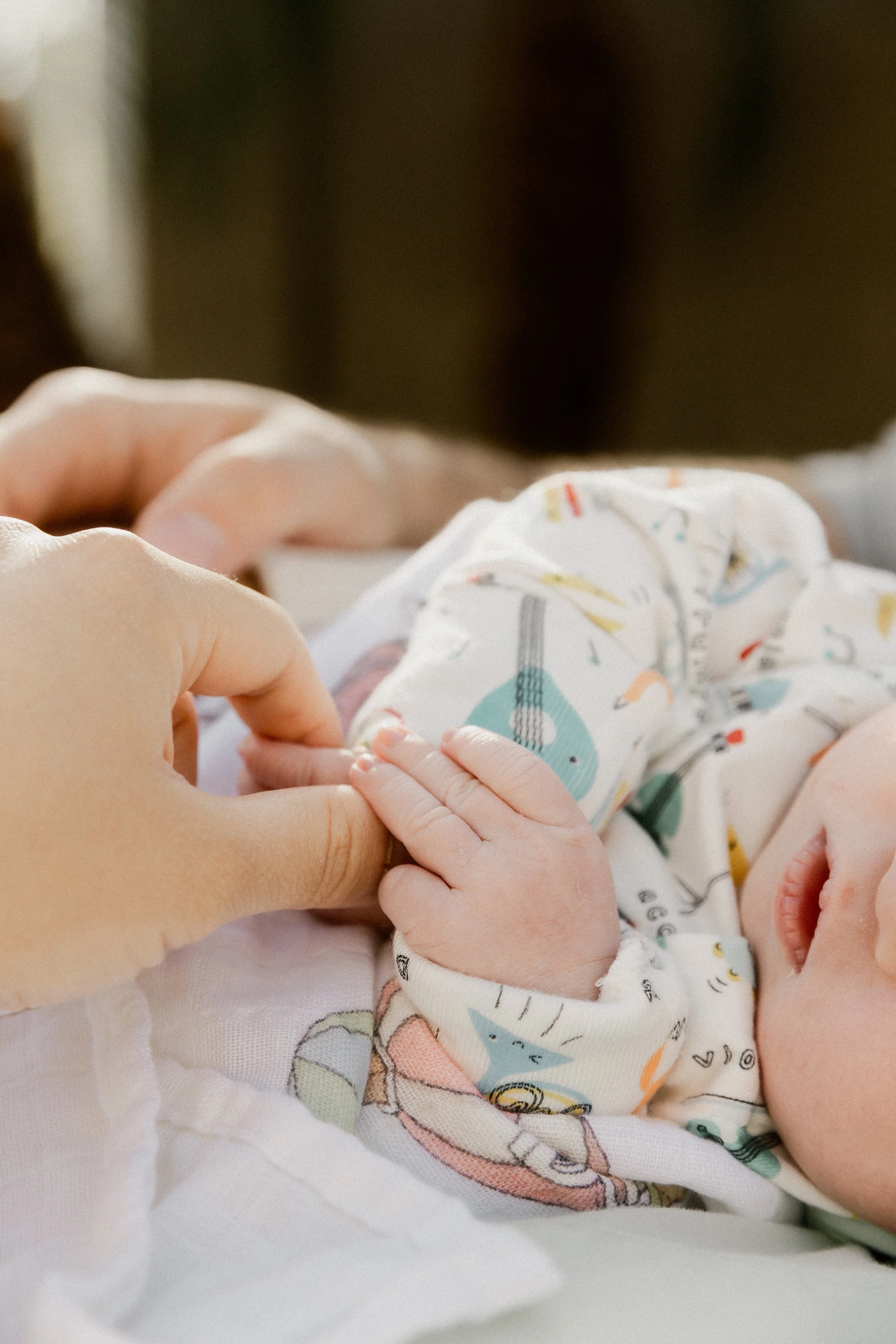A close-up of a baby's hand being held by an adult's hand, with the baby wearing a colorful, patterned onesie. The background is blurred.