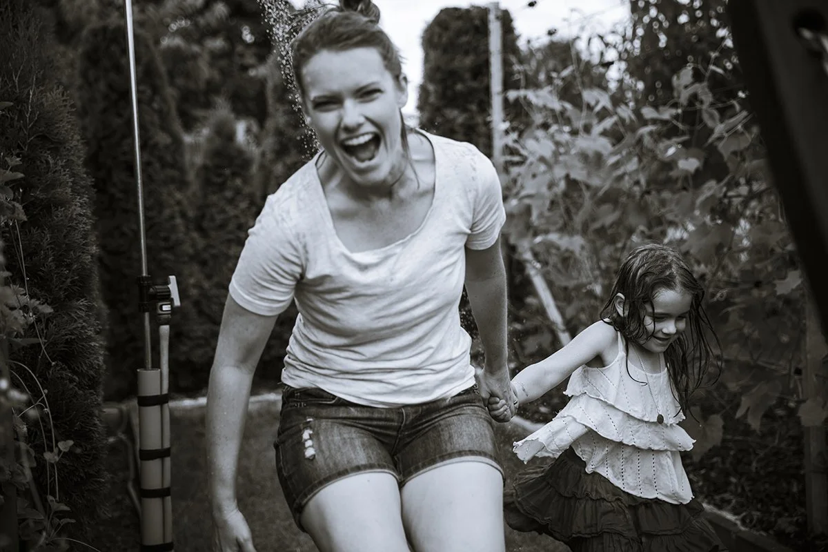 A woman and a young girl are playing outdoors, holding hands and smiling. The woman appears to be squatting or leaning forward, wearing a wet white t-shirt and denim shorts, with water splashing on her head. The girl has wet hair and is wearing a layered top and a ruffled skirt. They are in a garden or park with trees and bushes in the background.