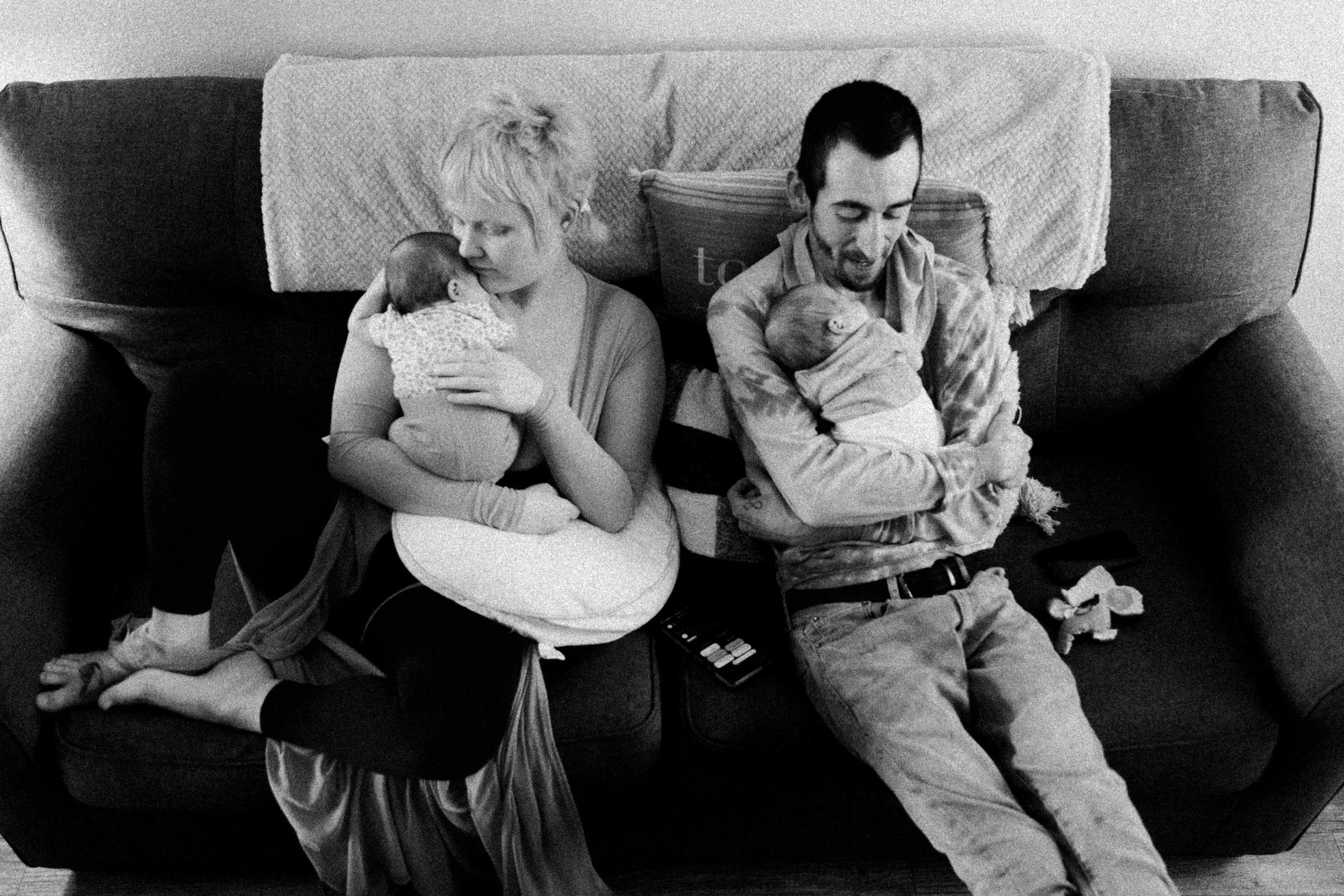A black and white photo of a woman and man sitting on a couch, each holding a baby. The woman is cuddling her baby close to her face, while the man is smiling and looking at his baby. There are pillows and a remote control on the couch, and some clot