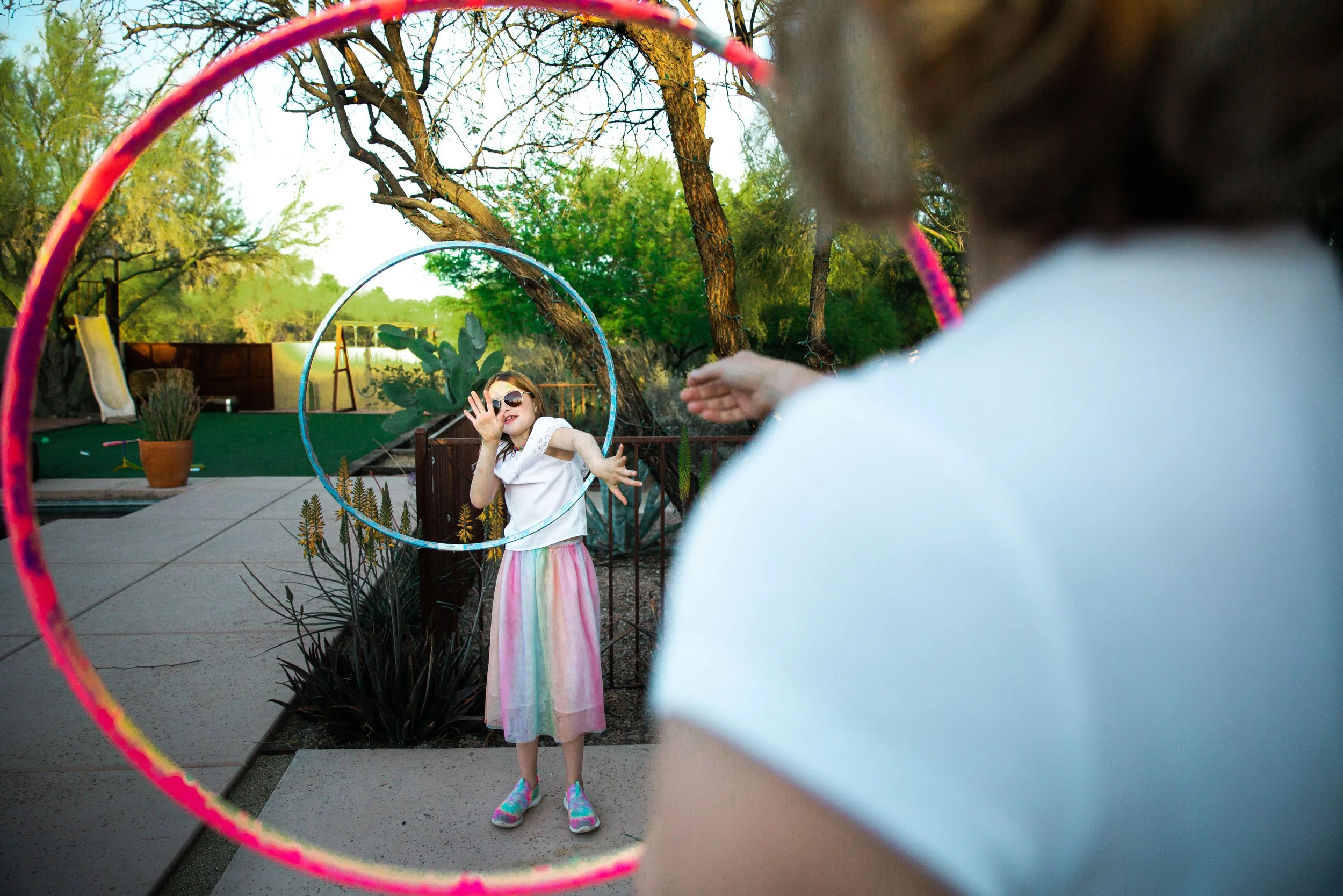 A young girl wearing sunglasses, a white top, and a colorful skirt is playing with hula hoops outdoors. The photo is taken through two overlapping hula hoops, one pink and one blue. The girl is smiling and reaching out with her left arm, with trees a