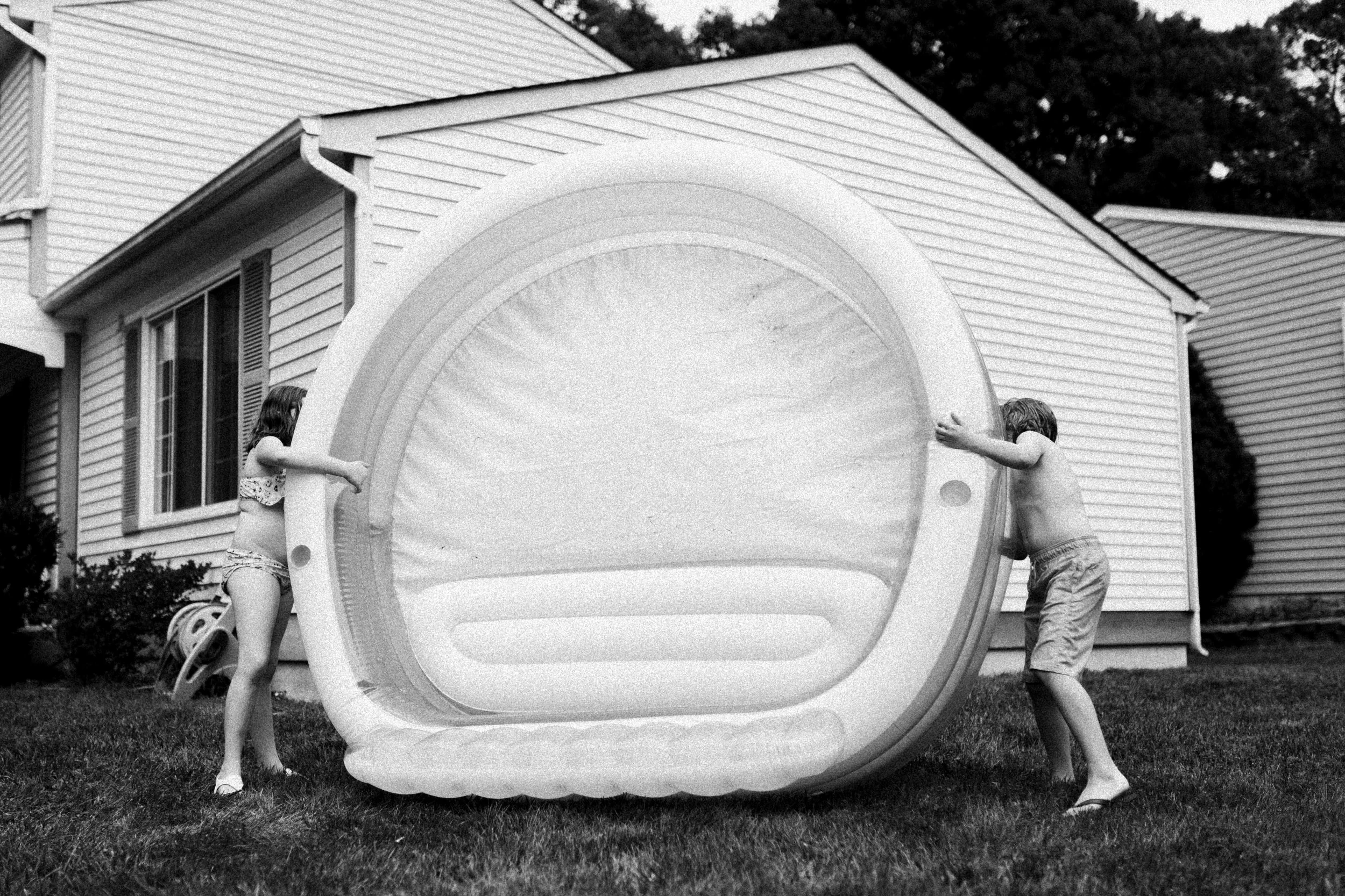 Two children, a girl wearing a swimsuit and a boy wearing shorts,  with an inflatable pool in a backyard. The girl is on one side and the boy on the other. There is a house with vinyl siding and a window in the background.