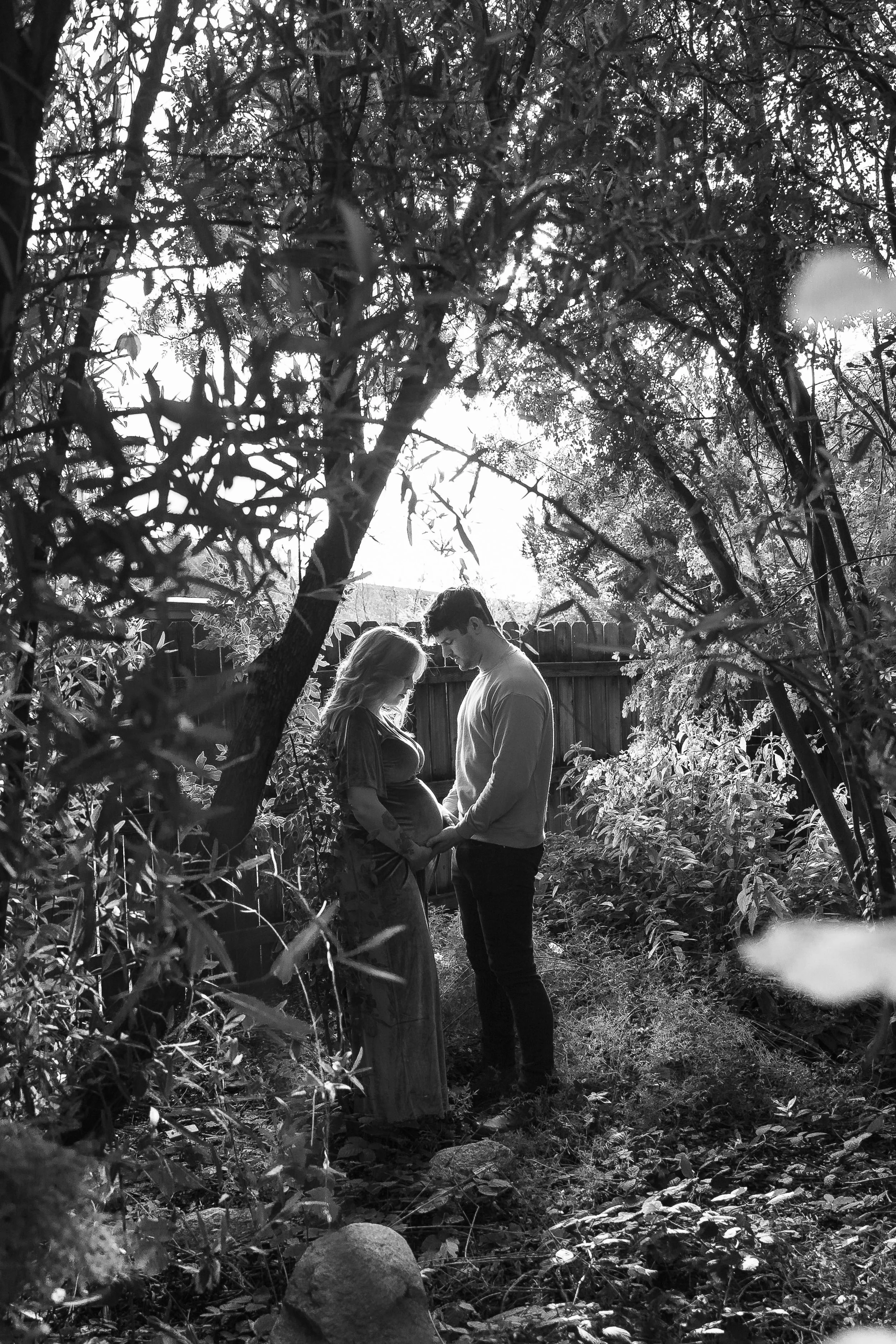 A black and white photo of a couple standing in a backyard, holding hands and smiling, surrounded by trees and plants, with a wooden fence in the background.