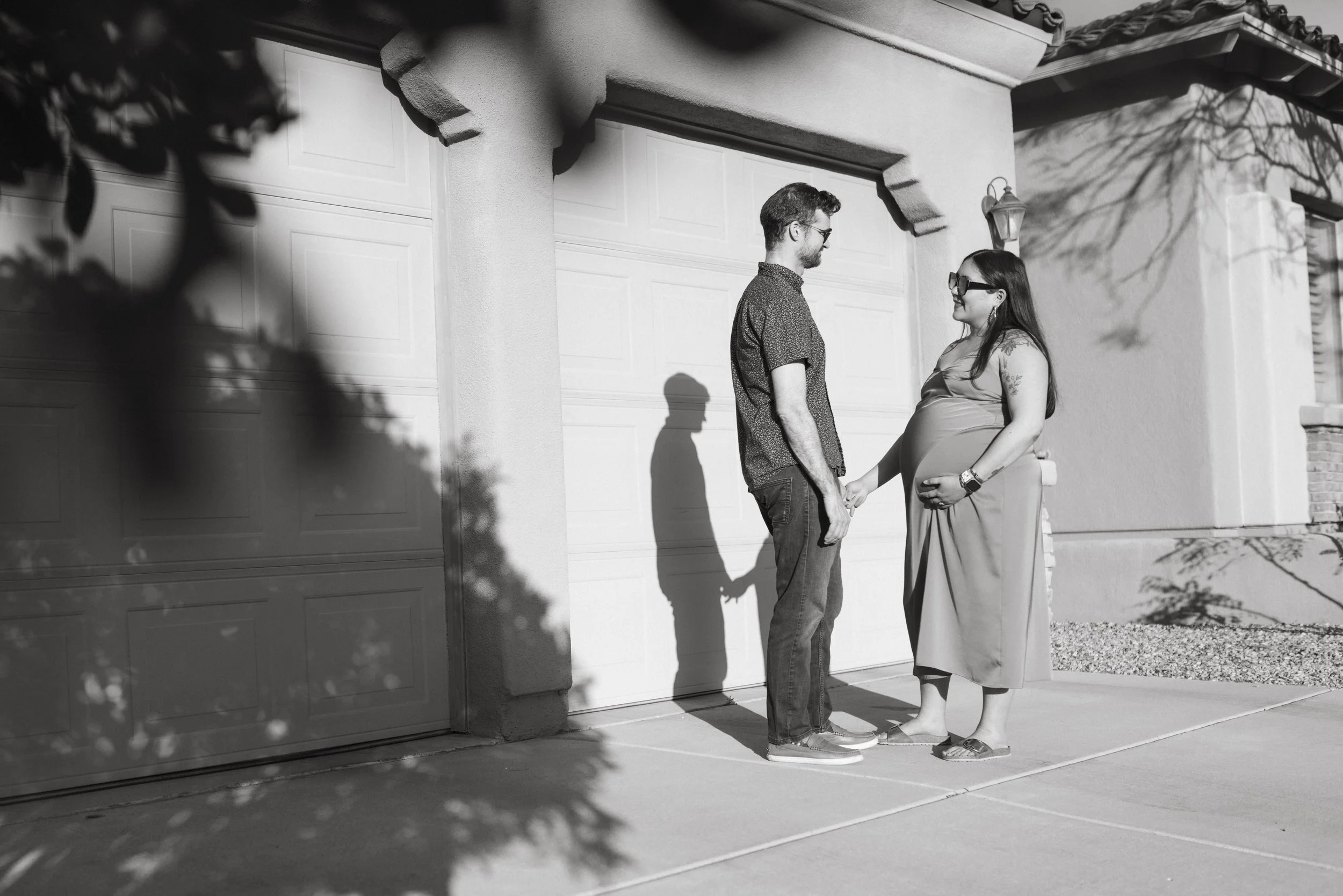 Black and white photo of a pregnant woman and a man holding hands outside a garage. They are looking at each other, with shadows cast on the wall.