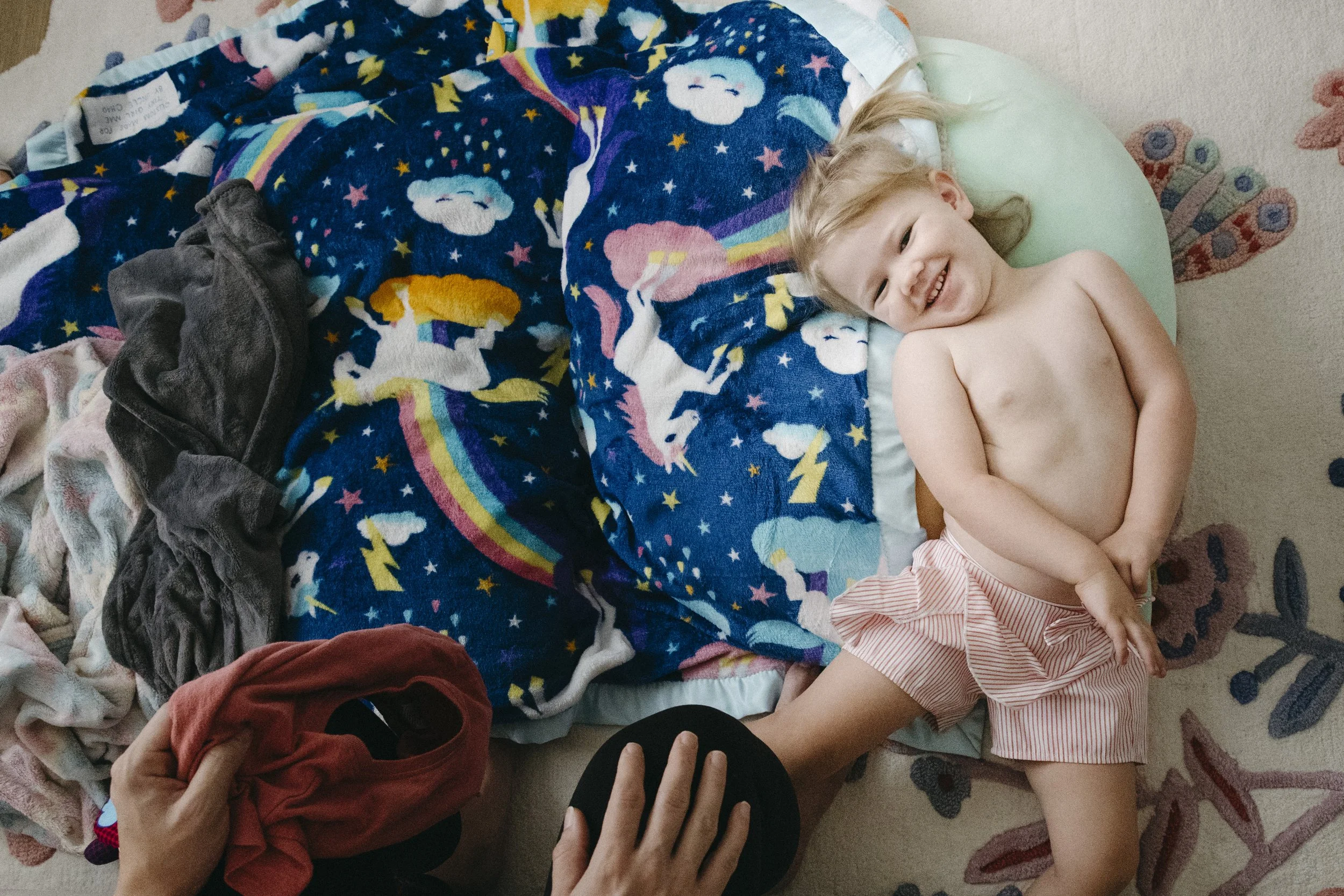 A young, shirtless child with blonde hair lying on a bed, smiling and wearing pink and white striped shorts. The bed has a colorful unicorn and rainbow themed blanket. Someone is holding a red shirt and a black sock or shoe, with their hand visible.
