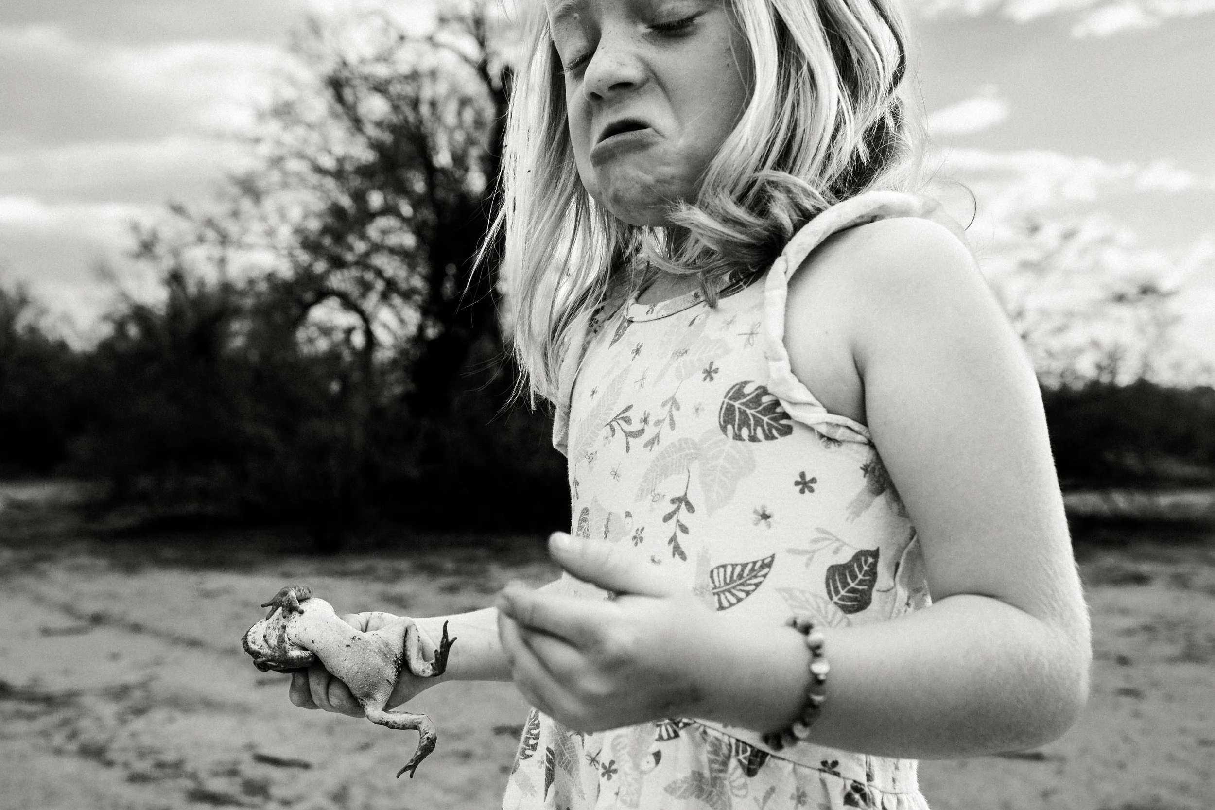 A young girl with light-colored hair, wearing a dress with leaf and plant patterns, holding a frog in her hands outdoors with trees and clouds in the background, in black and white.