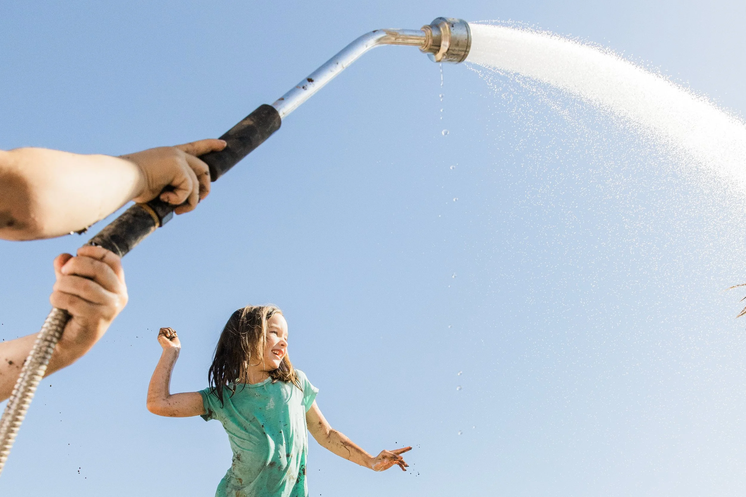 A young girl with long brown hair smiling and running through a hose shower on a sunny day.
