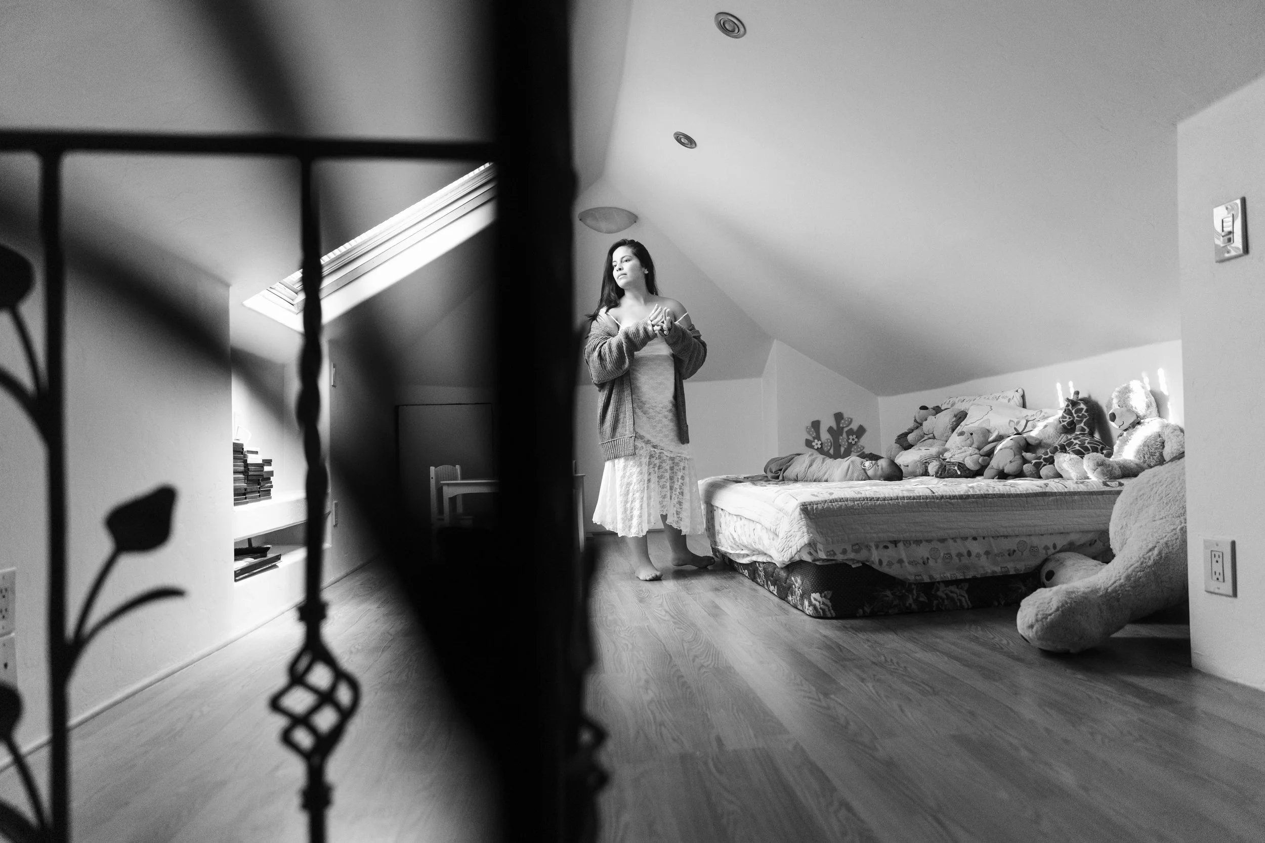 A black and white photo of a woman standing in a bedroom, seen through a decorative metal railing in the foreground. The woman has long dark hair, is wearing a cardigan and a long skirt, and looks contemplative. The room has a sloped ceiling with a s