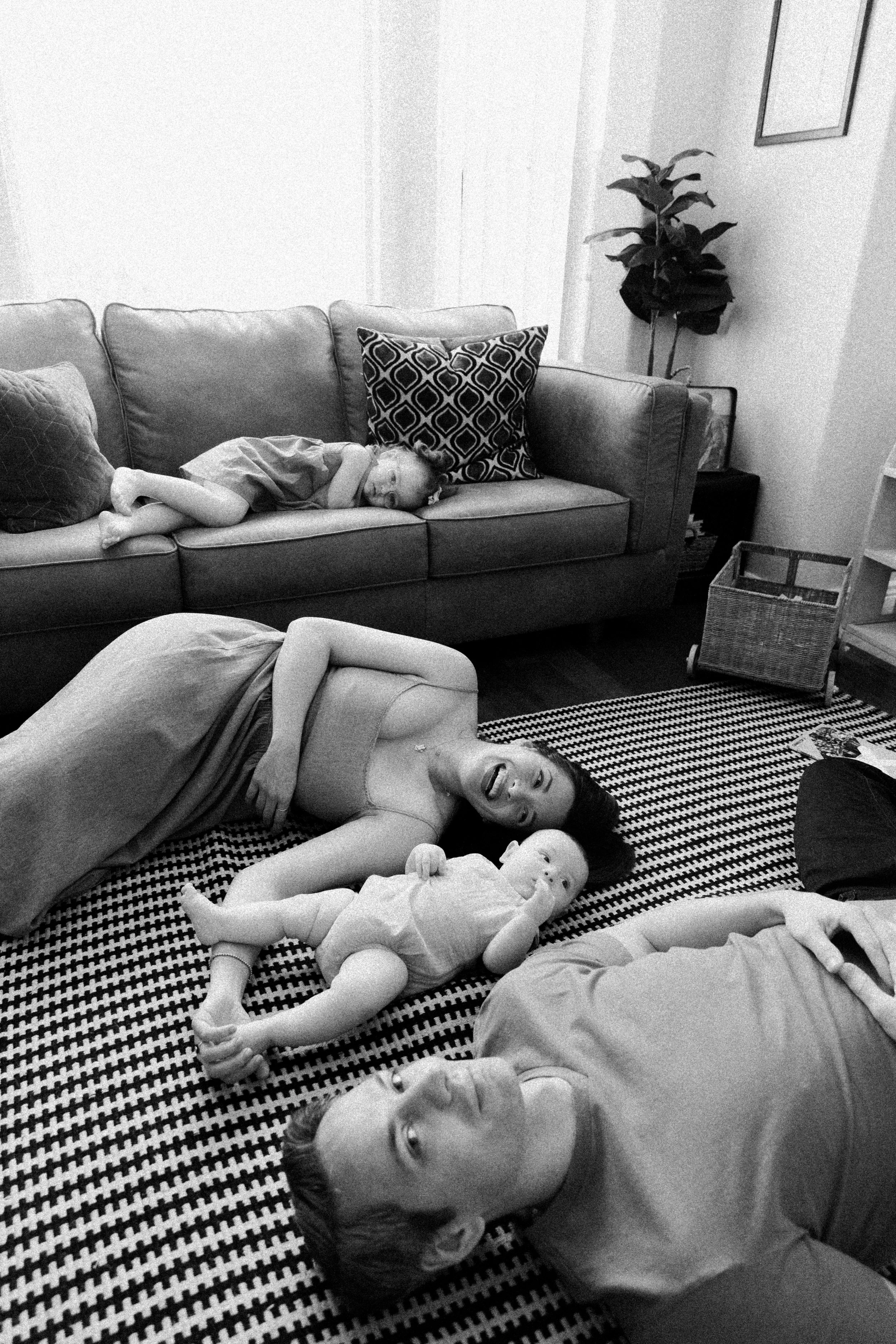 A woman, her husband and two children lying on the living room floor; one child nestled on the couch, the other awake and looking at the camera .