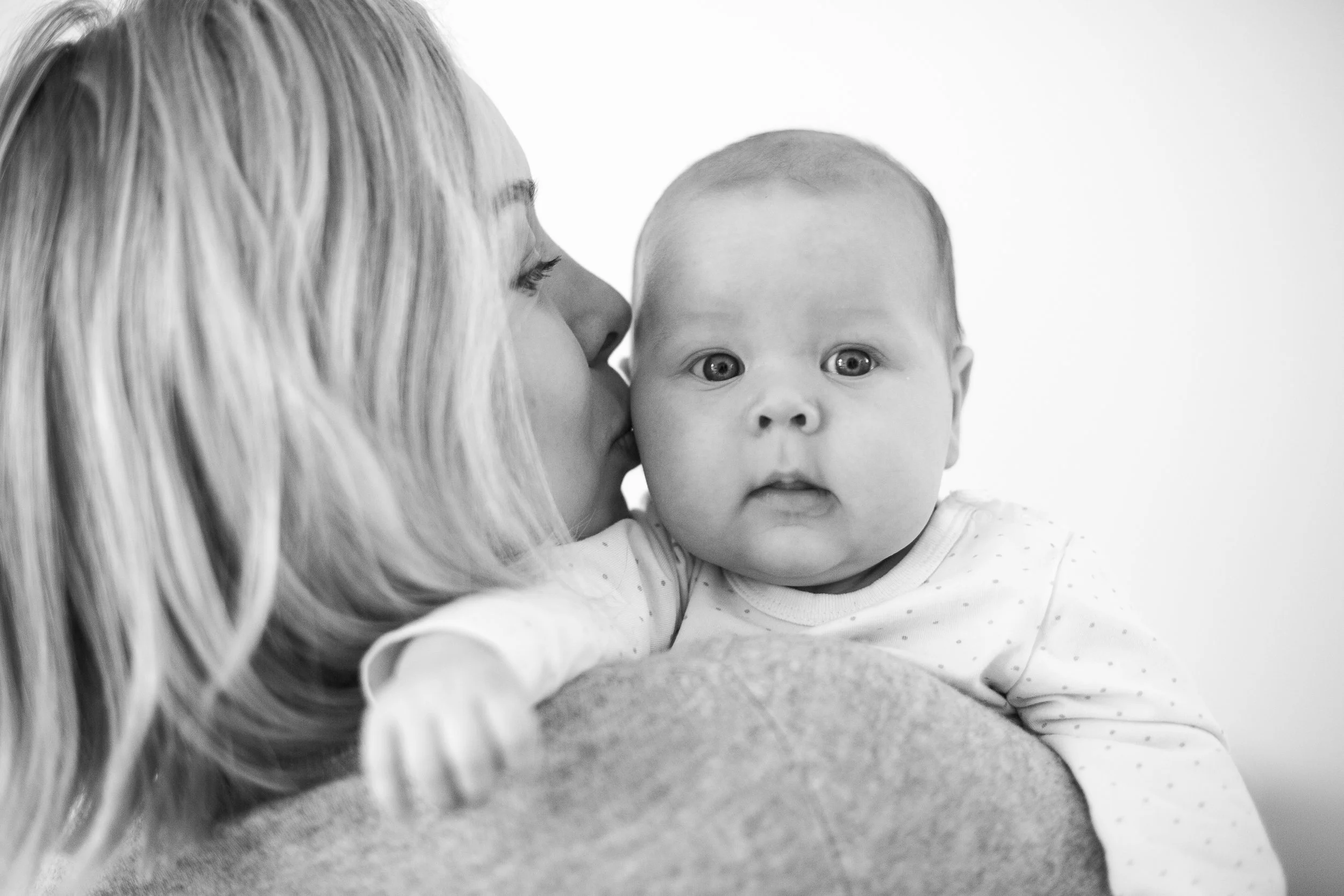 A woman with long blonde hair is kissing a baby on the cheek. The baby has light skin, big eyes, and is looking at the camera, wearing a white shirt with small polka dots. The photo is in black and white.