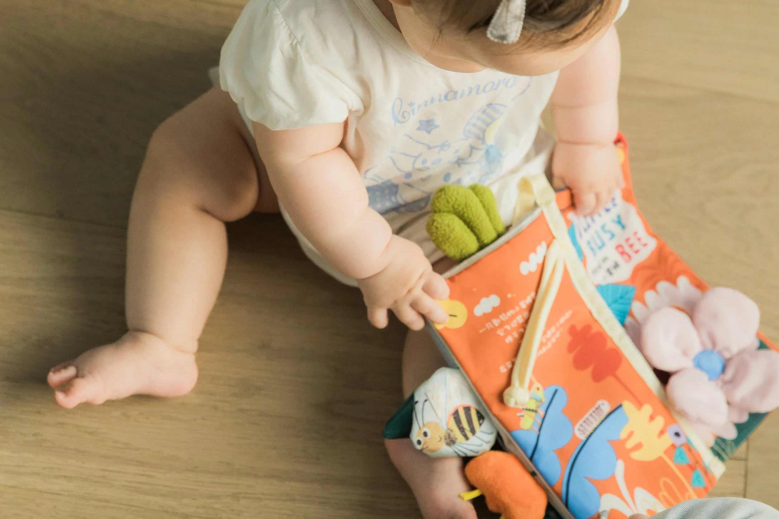 A baby sitting on a wooden floor, wearing a white shirt with blue writing, holding a green plush toy, with a colorful book nearby.