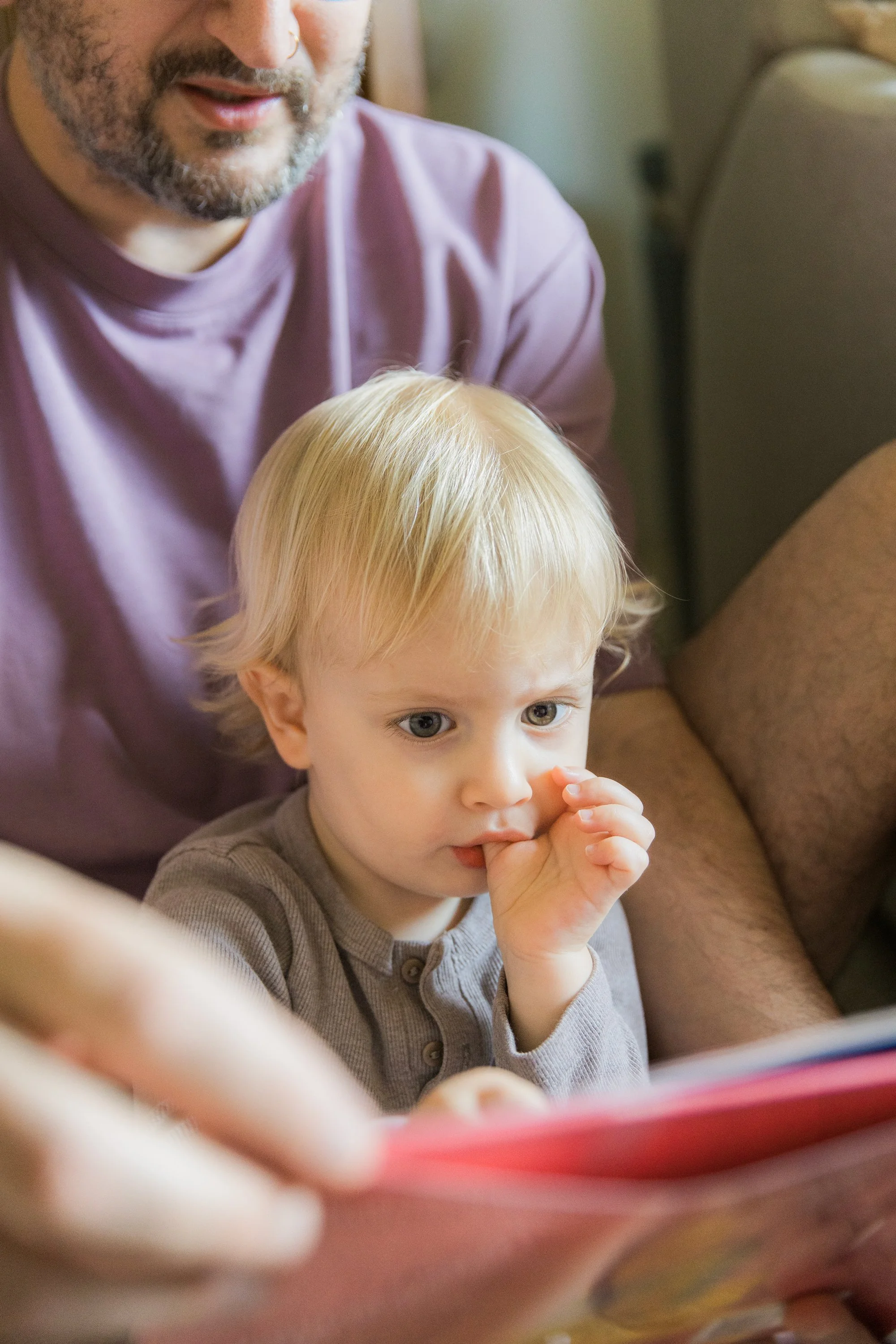 A young boy with blond hair and blue eyes sitting on a beige couch, looking at a book, with an adult man smiling beside him.