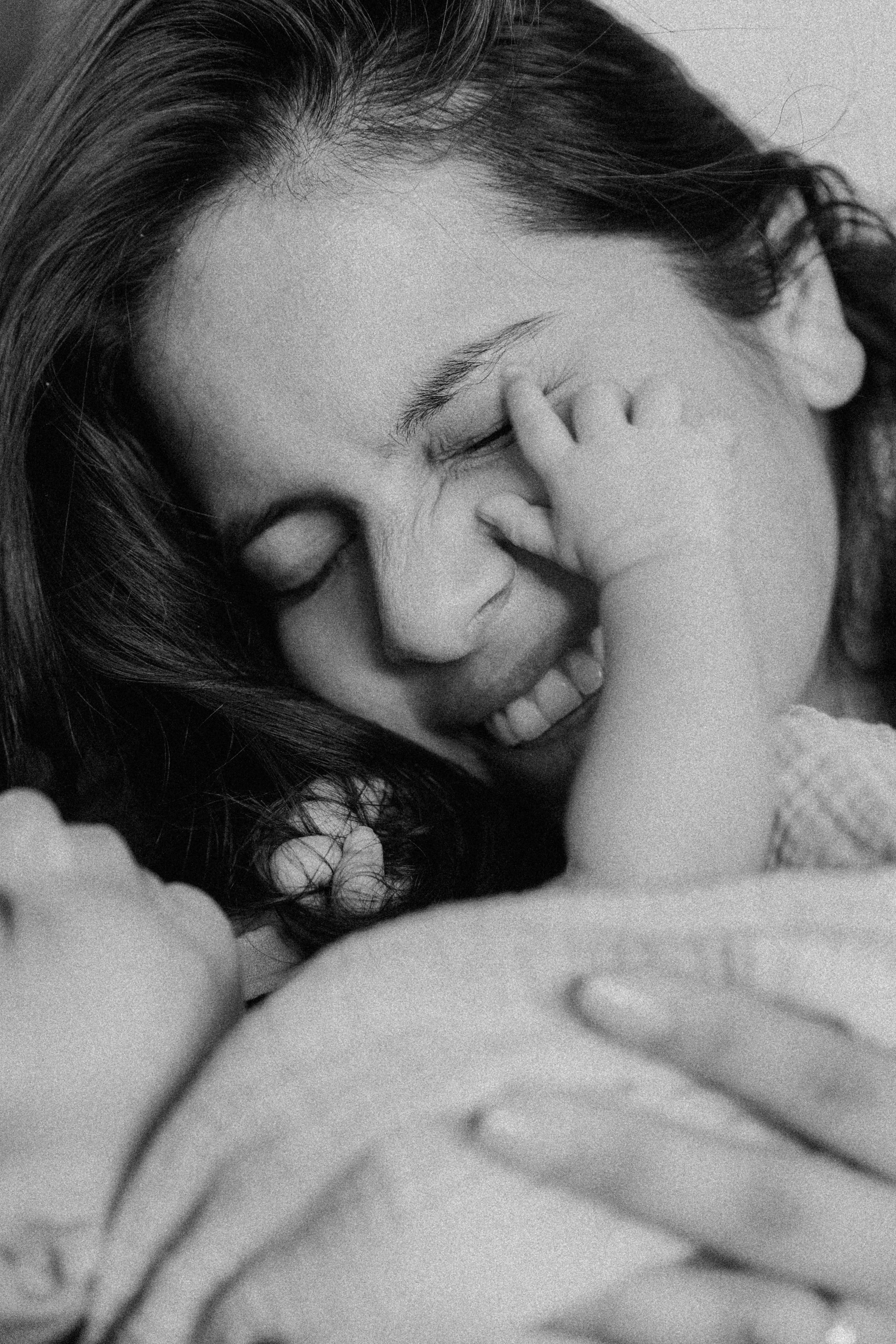 A woman with long dark hair smiling and closing her eyes, with a child's hand gently touching her face in an intimate close-up black and white photograph.