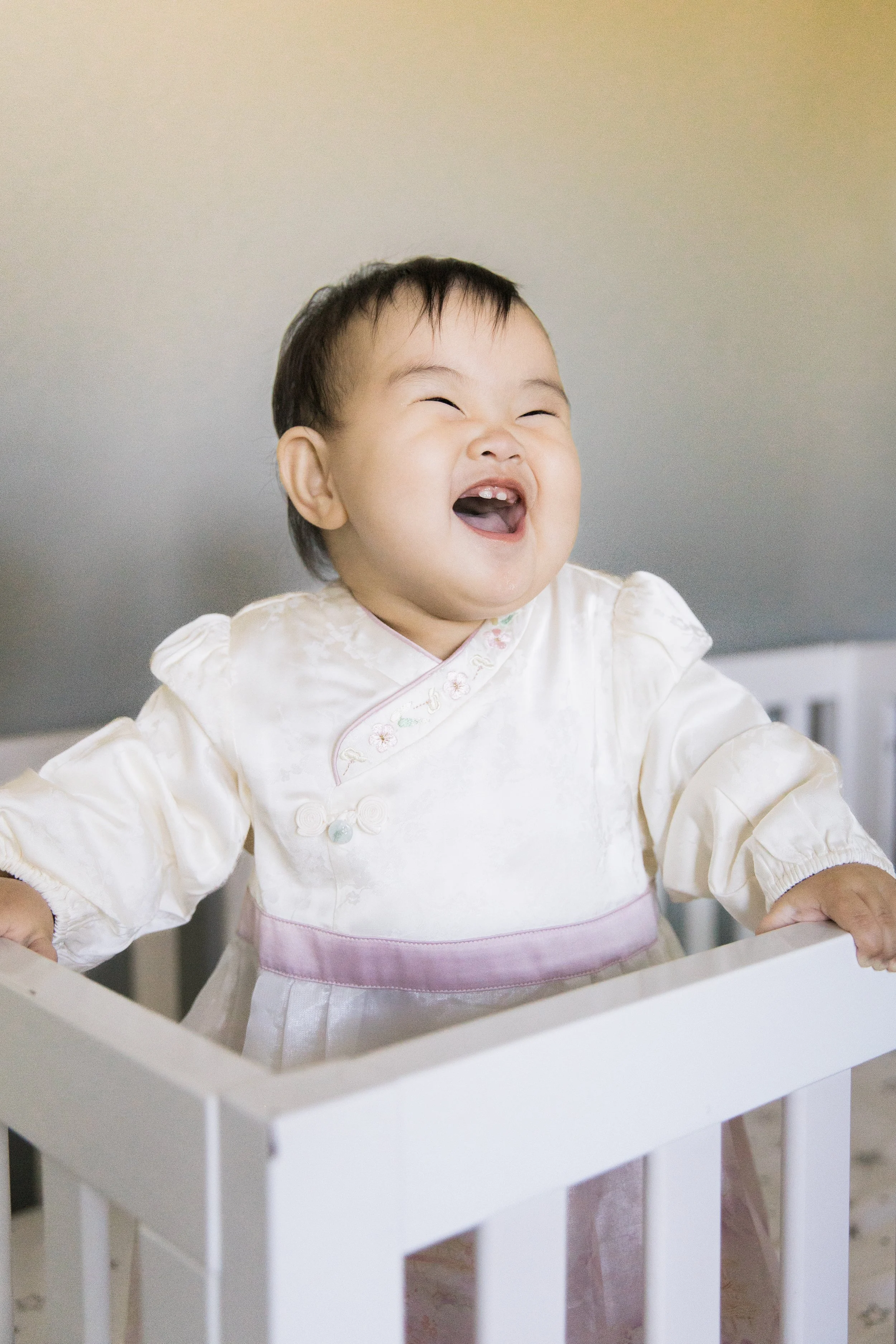 A young Asian child with short black hair, laughing with eyes closed, wearing a cream and pink traditional dress, standing behind a white wooden crib or playpen.