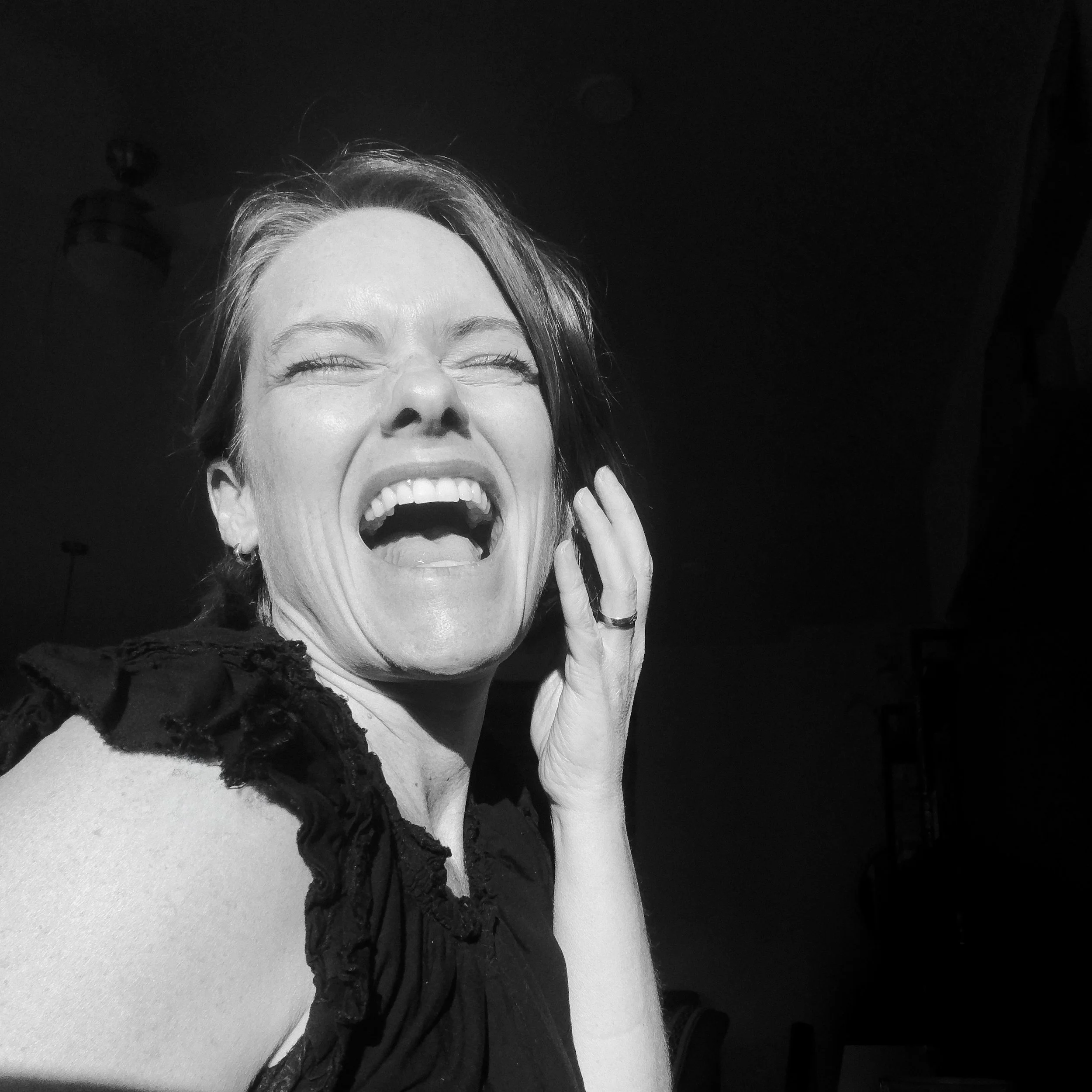 A woman laughing with eyes closed, hand touching cheek, wearing a black lace top, with a dark background and ceiling fan visible.