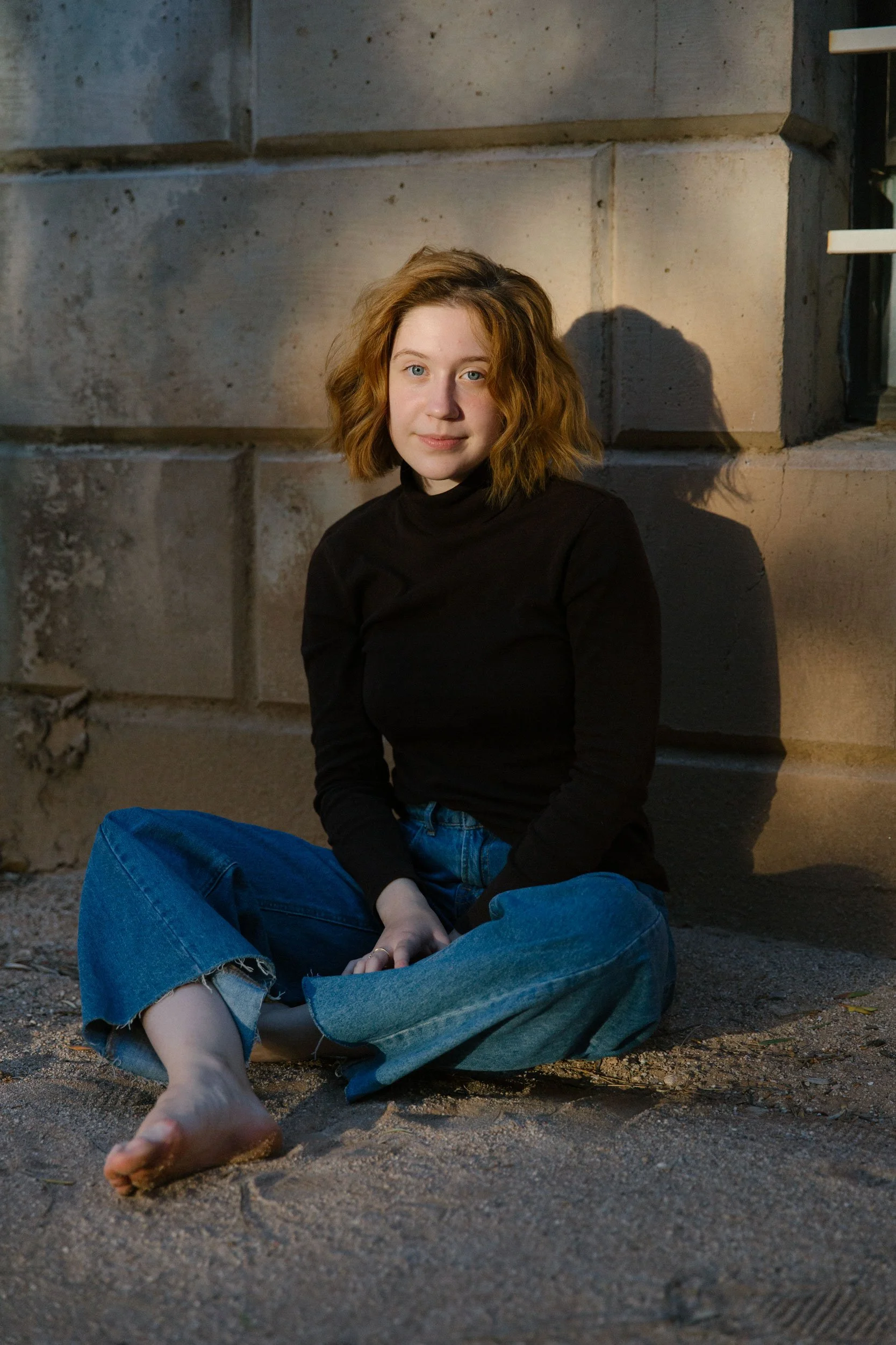 A young woman with red hair sitting on a sandy ground outdoors against a concrete wall, wearing a black turtleneck and blue jeans, looking at the camera.