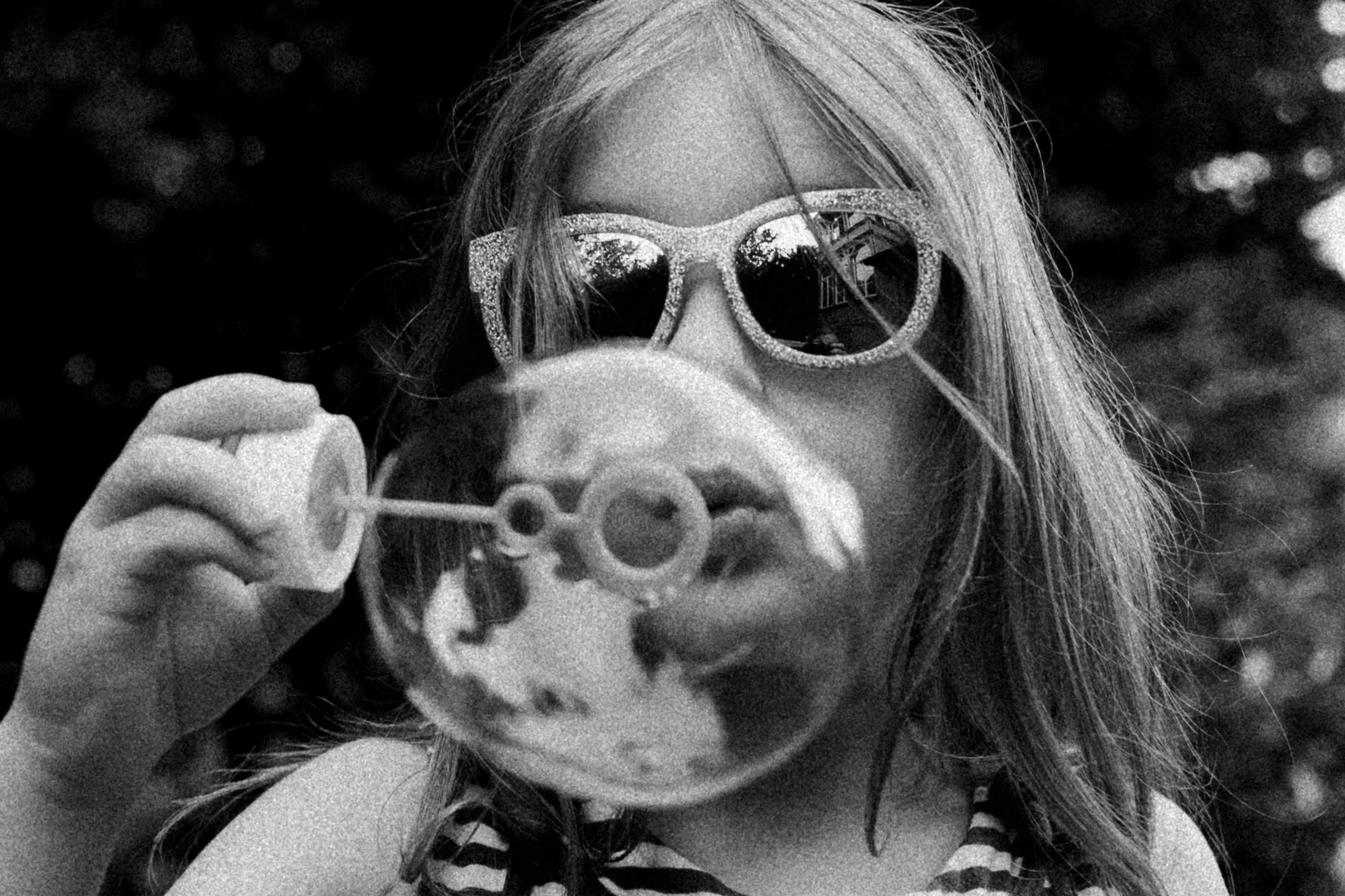 A girl with long hair wearing sunglasses with a glittery frame and striped clothing, blowing bubbles outdoors.