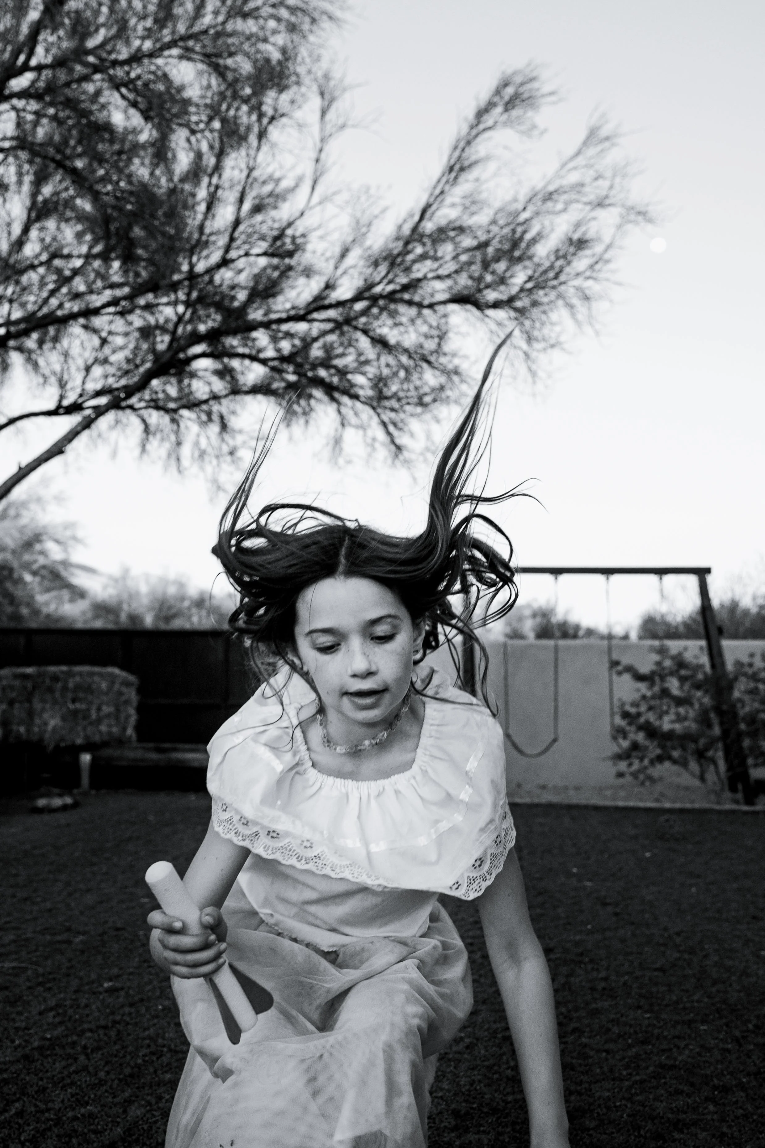 A girl in a white dress is running or jumping outdoors on a grassy yard, holding a toy in her hand, with her hair flying in the air, and trees and a swing set in the background.