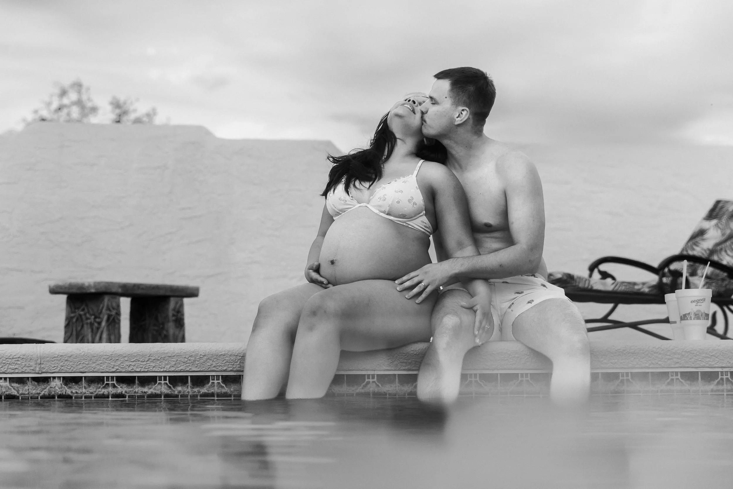 A pregnant woman and a man sitting by a pool, sharing a kiss. The woman is wearing a patterned bikini top and the man is shirtless, both are sitting on the pool's edge with their legs in the water. In the background, there are lounge chairs and a table, with cloudy skies.