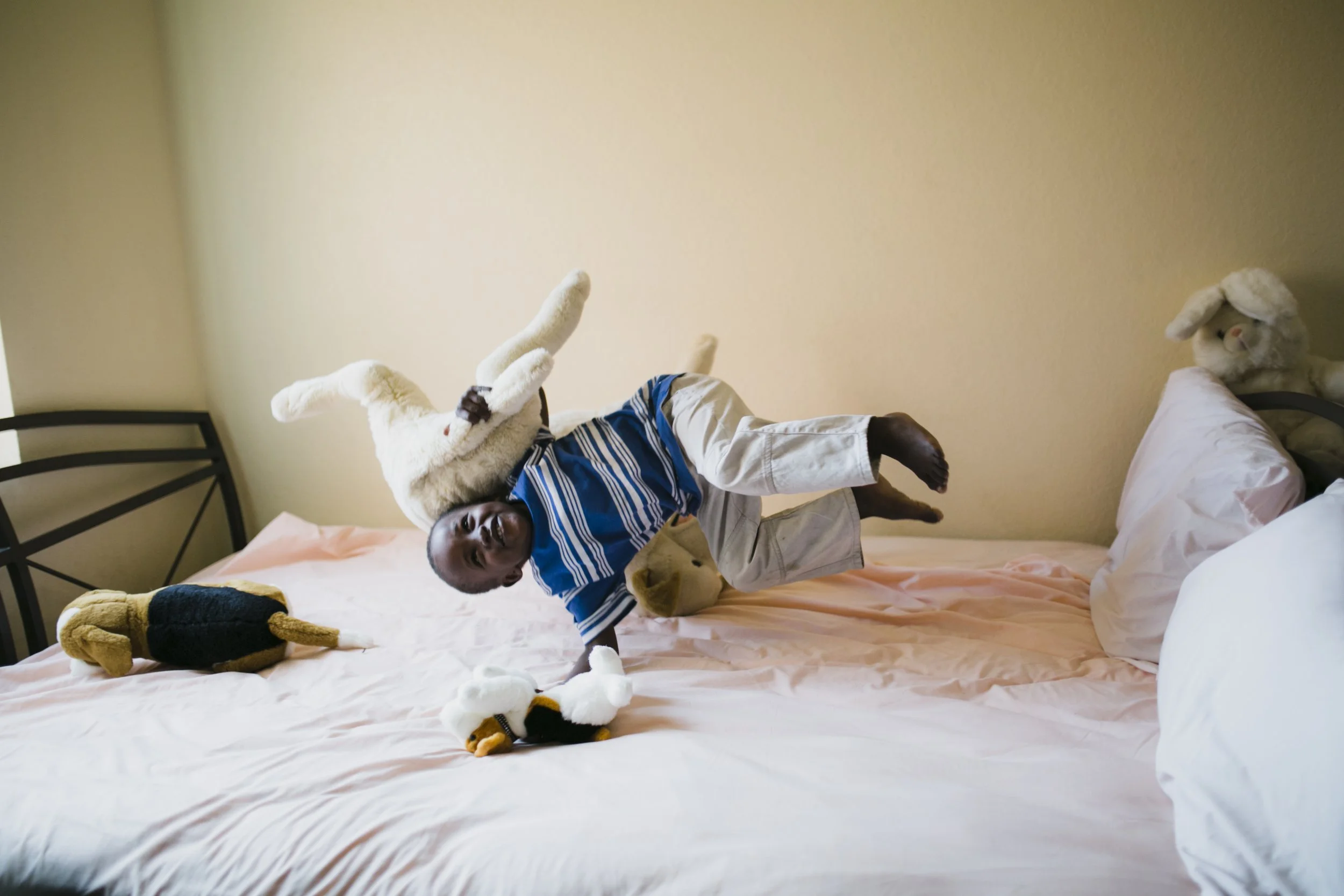 A young boy with dark skin playing on a bed with multiple stuffed animals, including a dog and a tiger. The boy is smiling and wearing a blue and white striped shirt with white shorts. The bed has a pinkish sheet and a sign of a pillow. The room has cream-colored walls and a black metal headboard.
