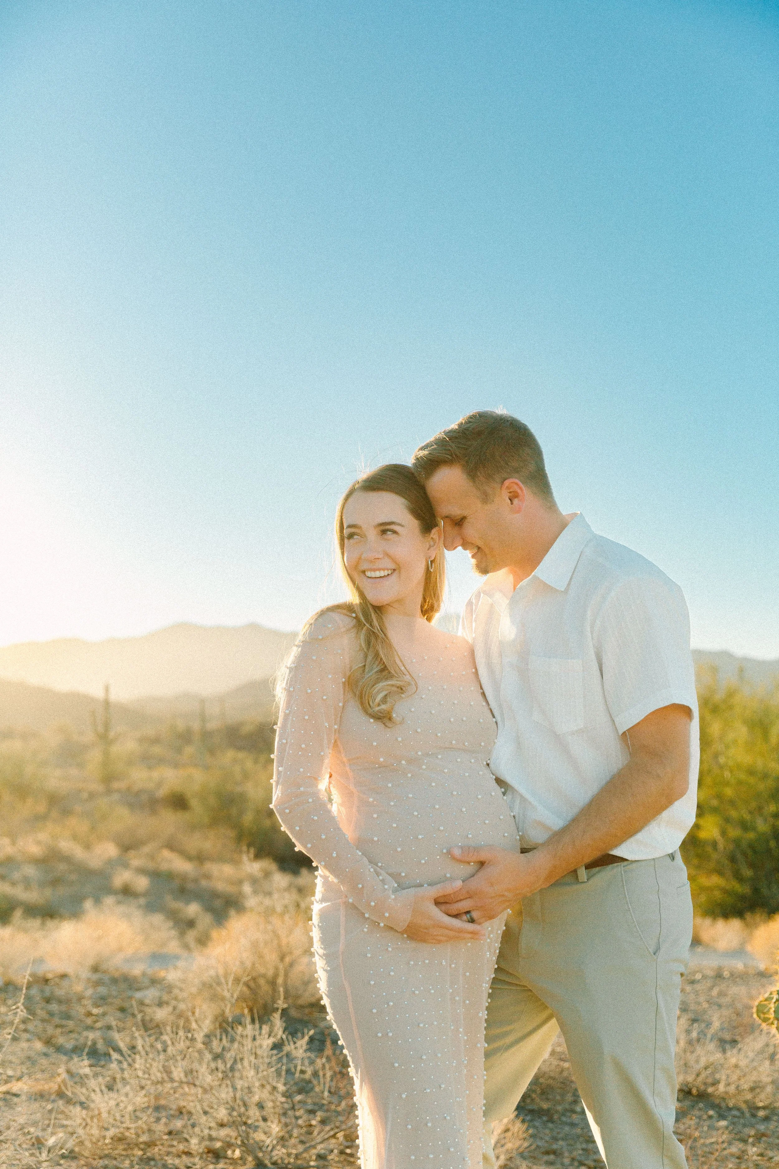 A smiling woman wearing a light dress and a man in light-colored clothing, standing outside in a desert or dry landscape during sunset, intertwined and sharing a moment of happiness.