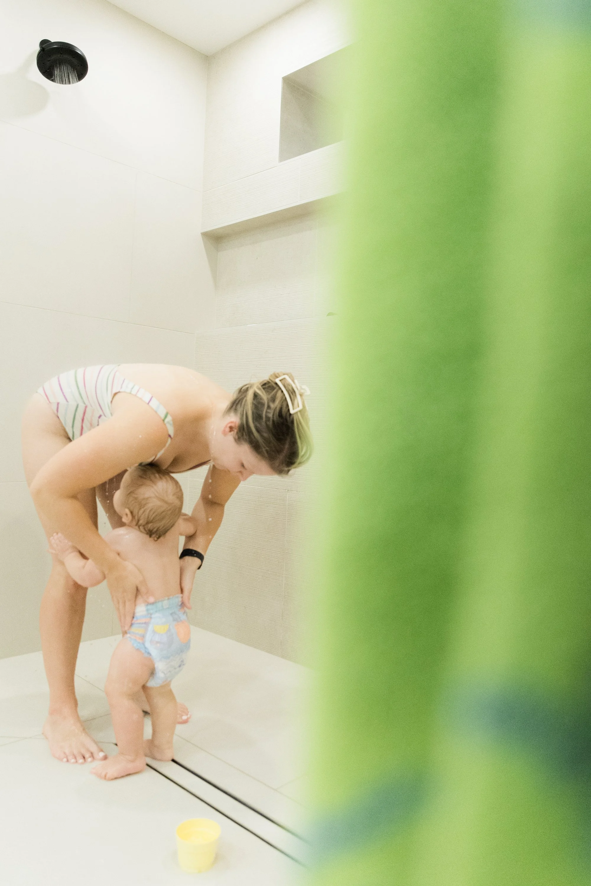 A woman wearing a striped shirt and glasses is helping a young child in a diaper in a shower. The woman is leaning over and supporting the child's body while the child is standing on the tile floor. A yellow container is on the floor nearby, and the 