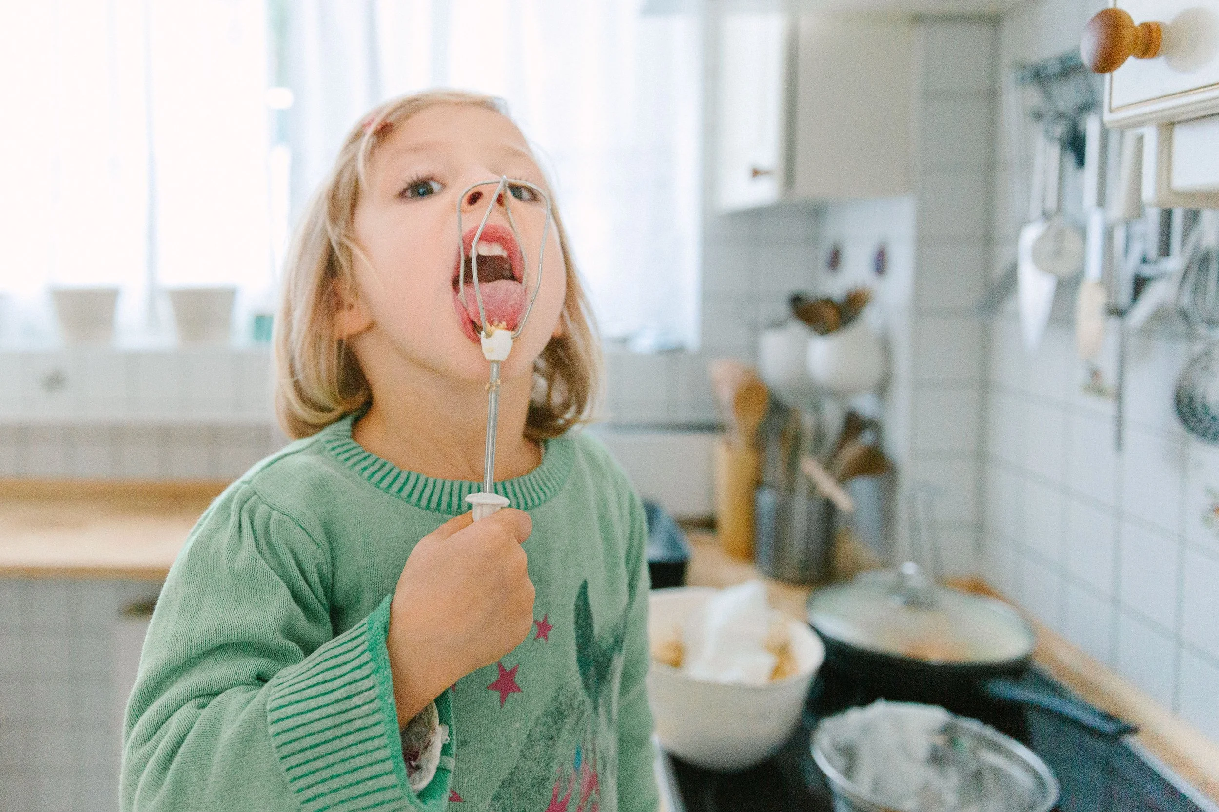 A young girl with blonde hair, wearing a green sweater with pink stars, holds a mouth guard and sticks her tongue out in a bright, clean kitchen with white tiles and shelves in the background.