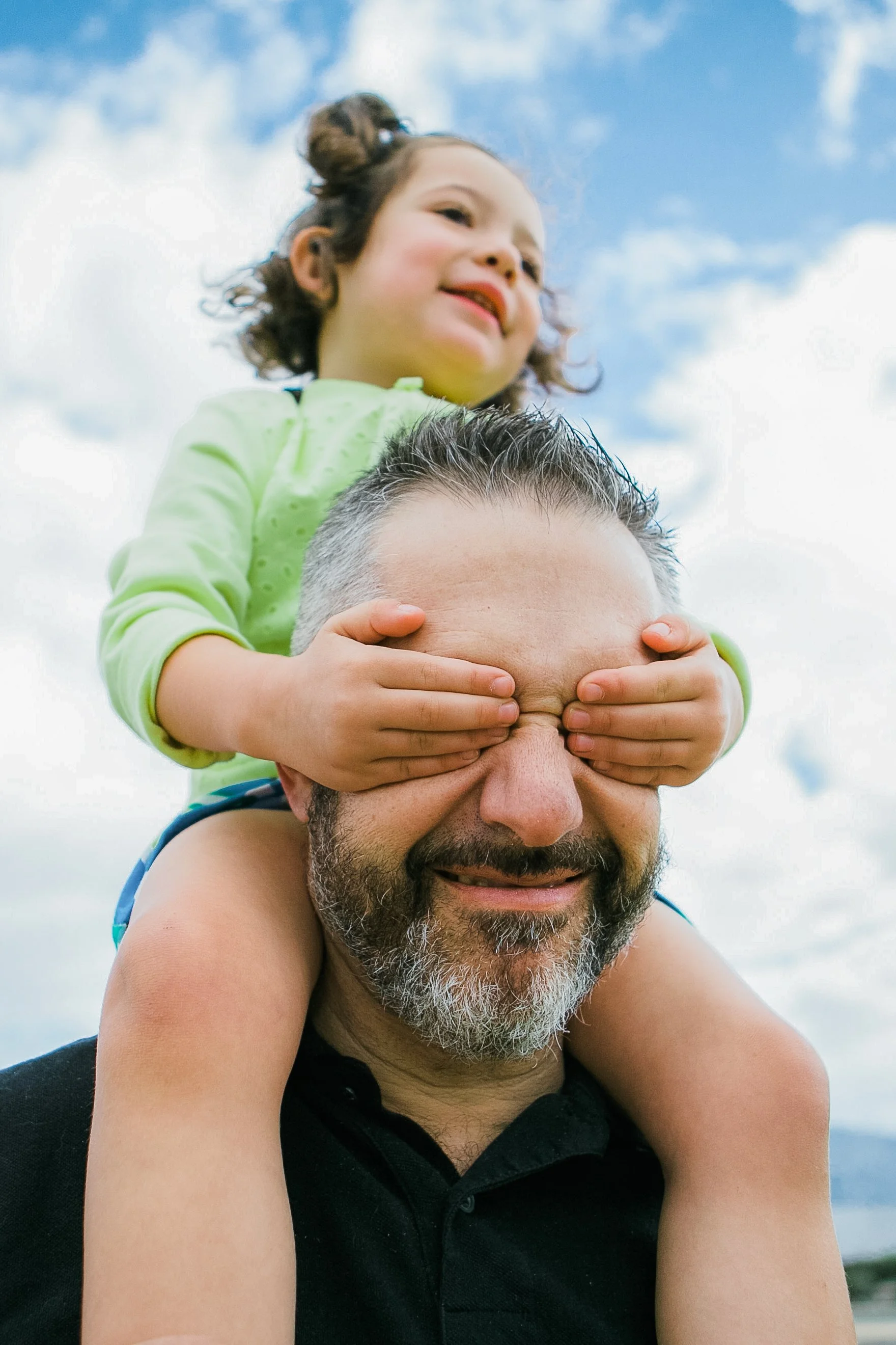 A man with gray hair and a beard giving a piggyback ride to a young girl with curly hair and a green jacket outdoors against a cloudy sky.