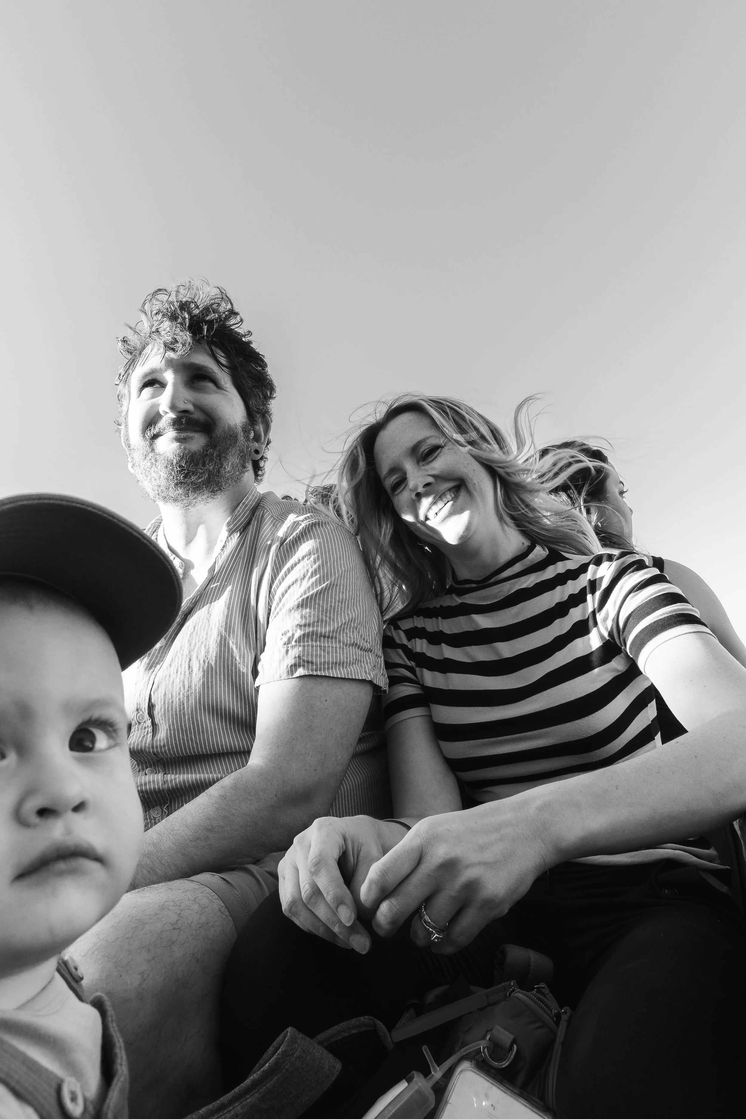 A black-and-white photo of a group of people, including a man with curly hair and a beard, a woman with long wavy hair, and a young child in the foreground, all smiling and looking in different directions, against a plain sky background.