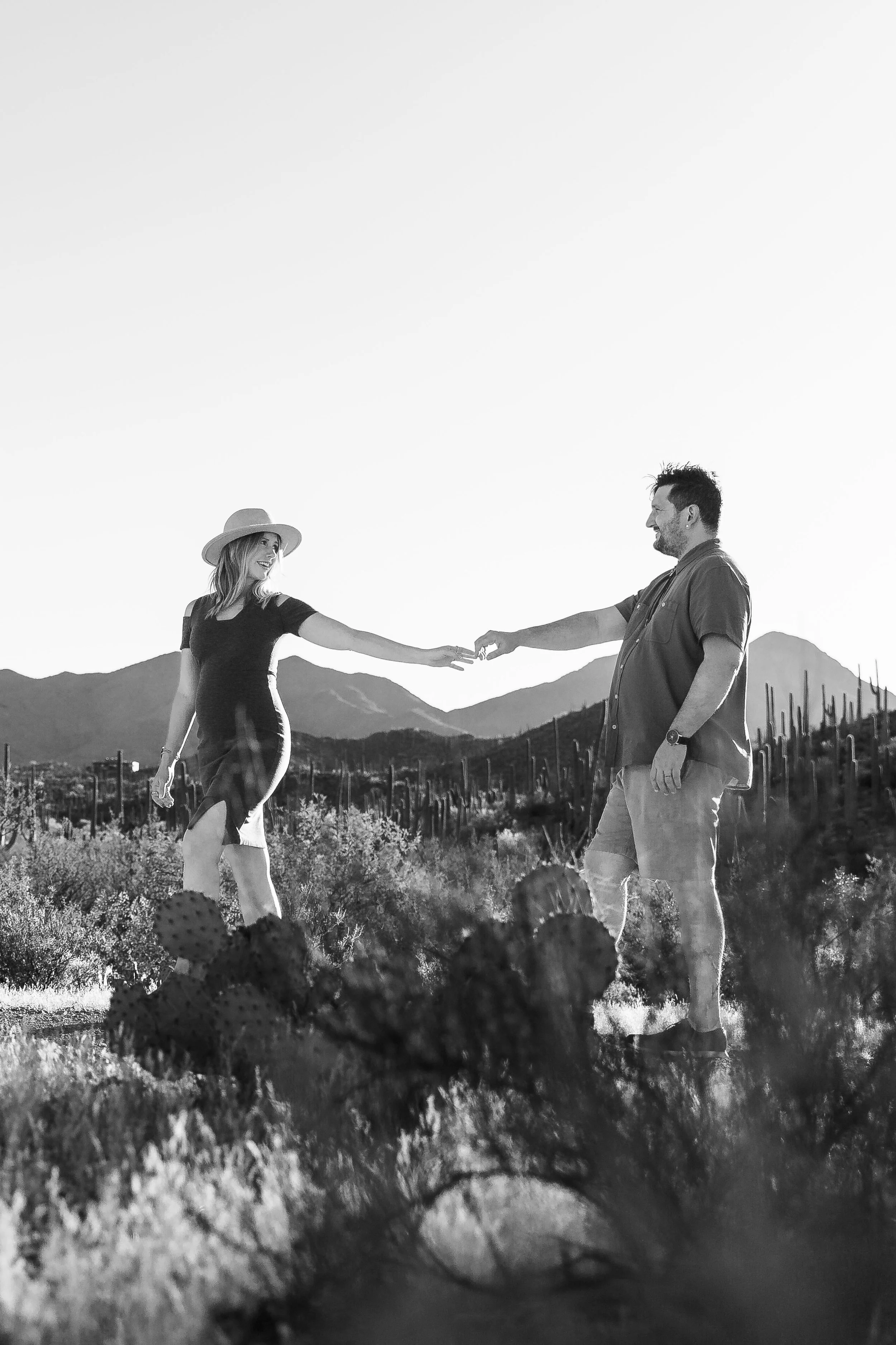 A black-and-white photo of a couple in a desert landscape. The woman, wearing a hat and a dress, is reaching out to the man, who is smiling and wearing a short-sleeved button-up shirt and shorts. Mountains and cacti are visible in the background.