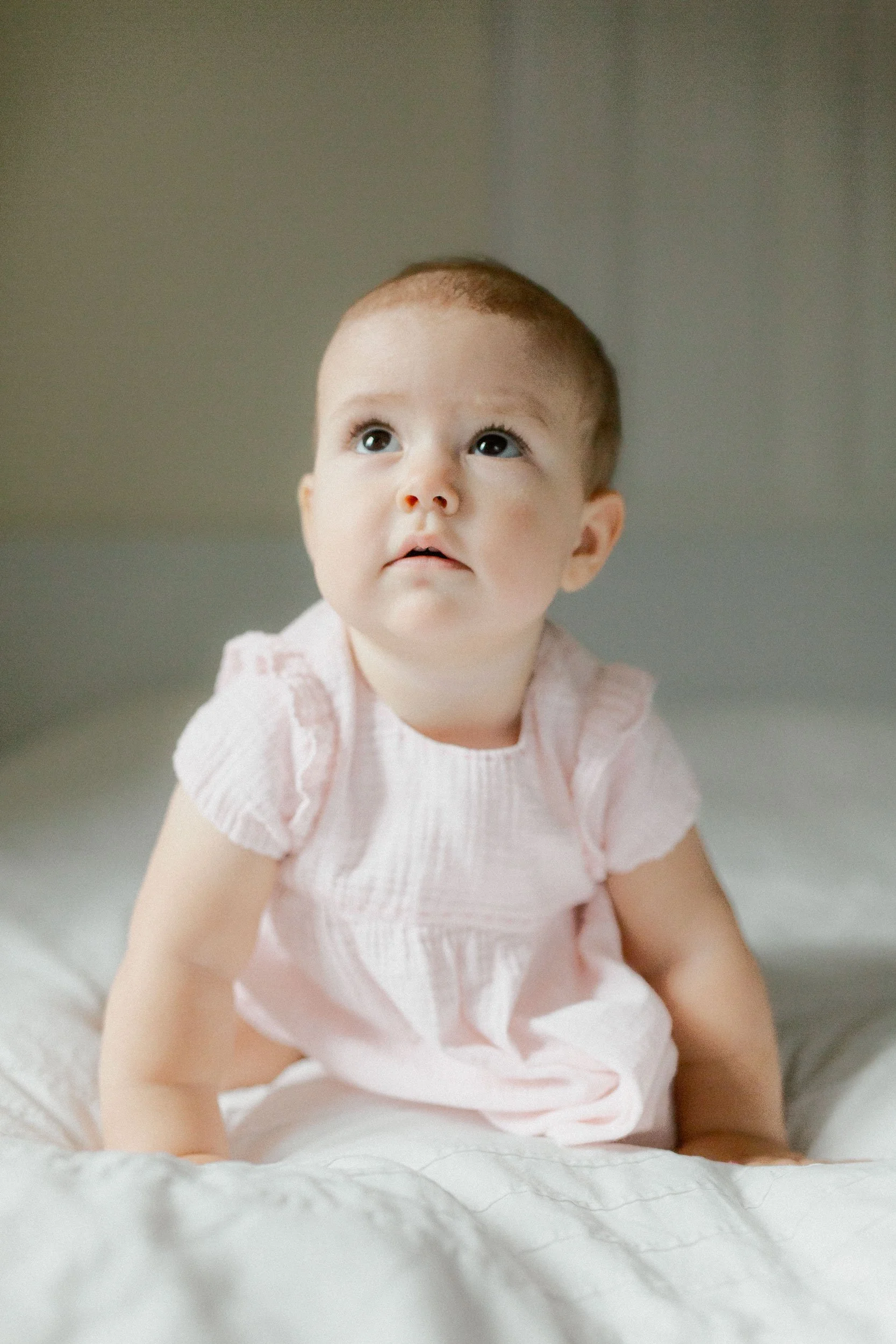 A young child with short brown hair and big eyes, wearing a light pink dress, sitting on a bed with cream-colored bedding, looking slightly upward.
