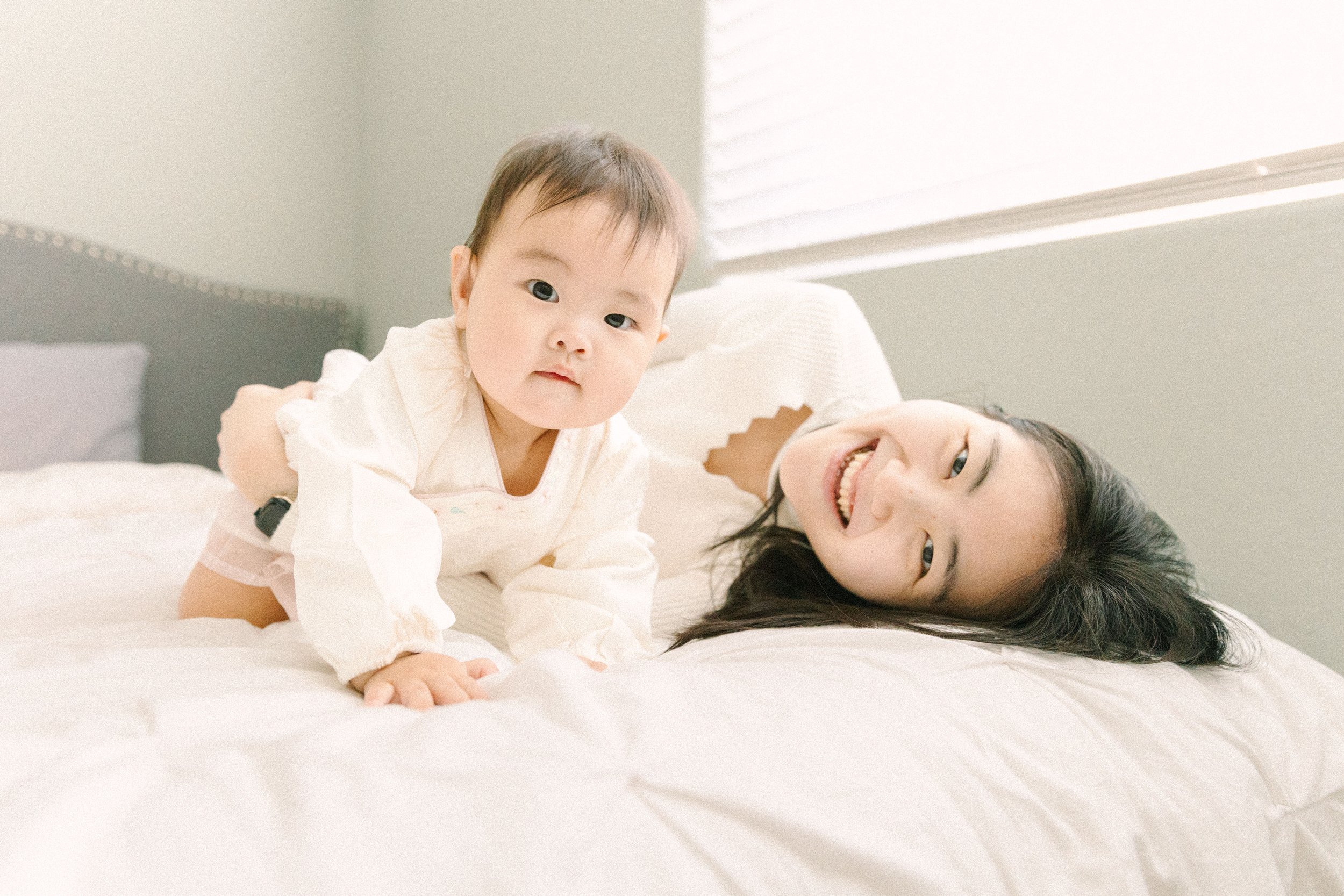 A young child and an adult woman lying on a bed, smiling and looking at the camera, with sunlight coming through the window.