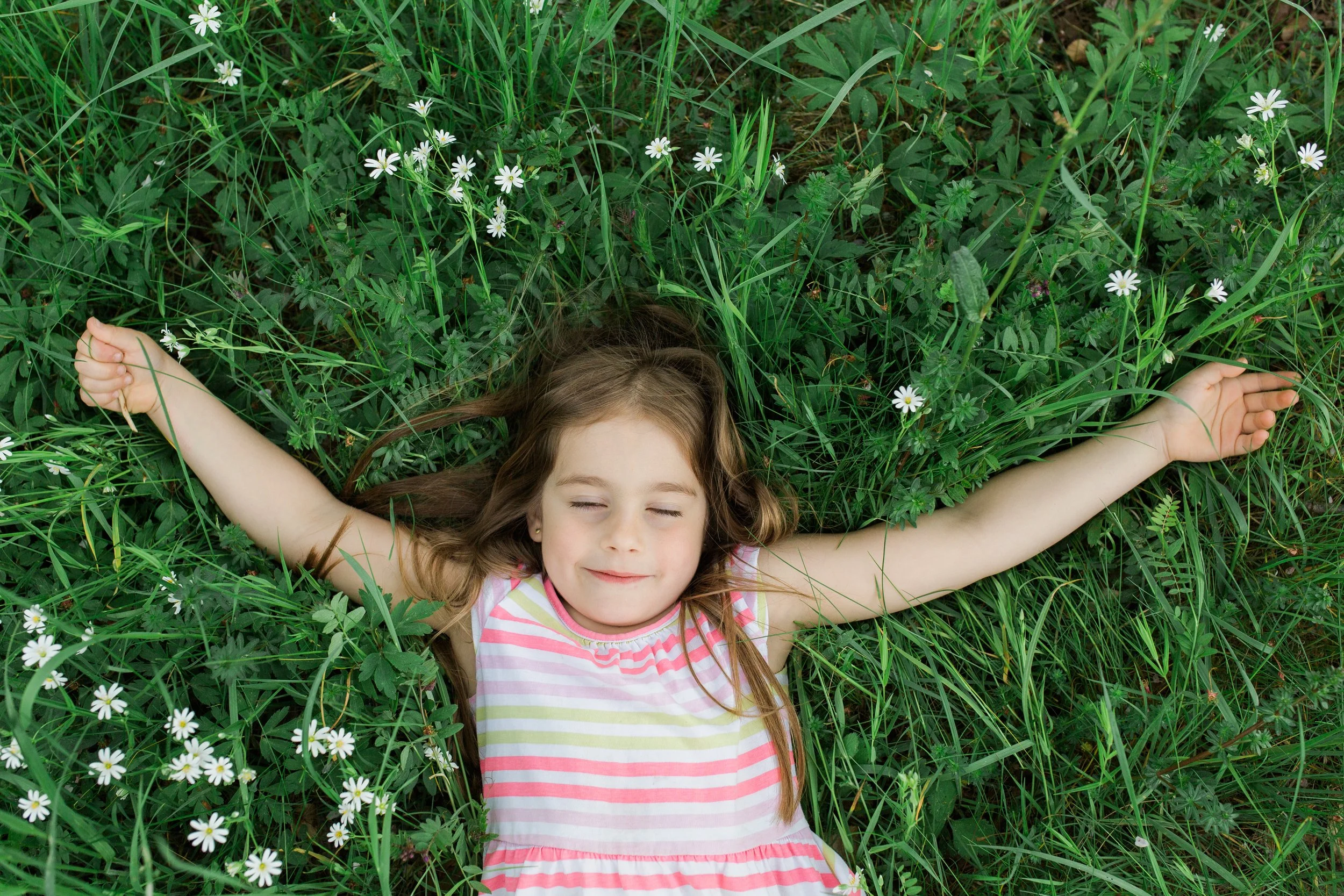 Young girl with long brown hair, wearing a pink and white striped dress, lying on her back among green grass and white daisies, with her eyes closed and arms spread out.