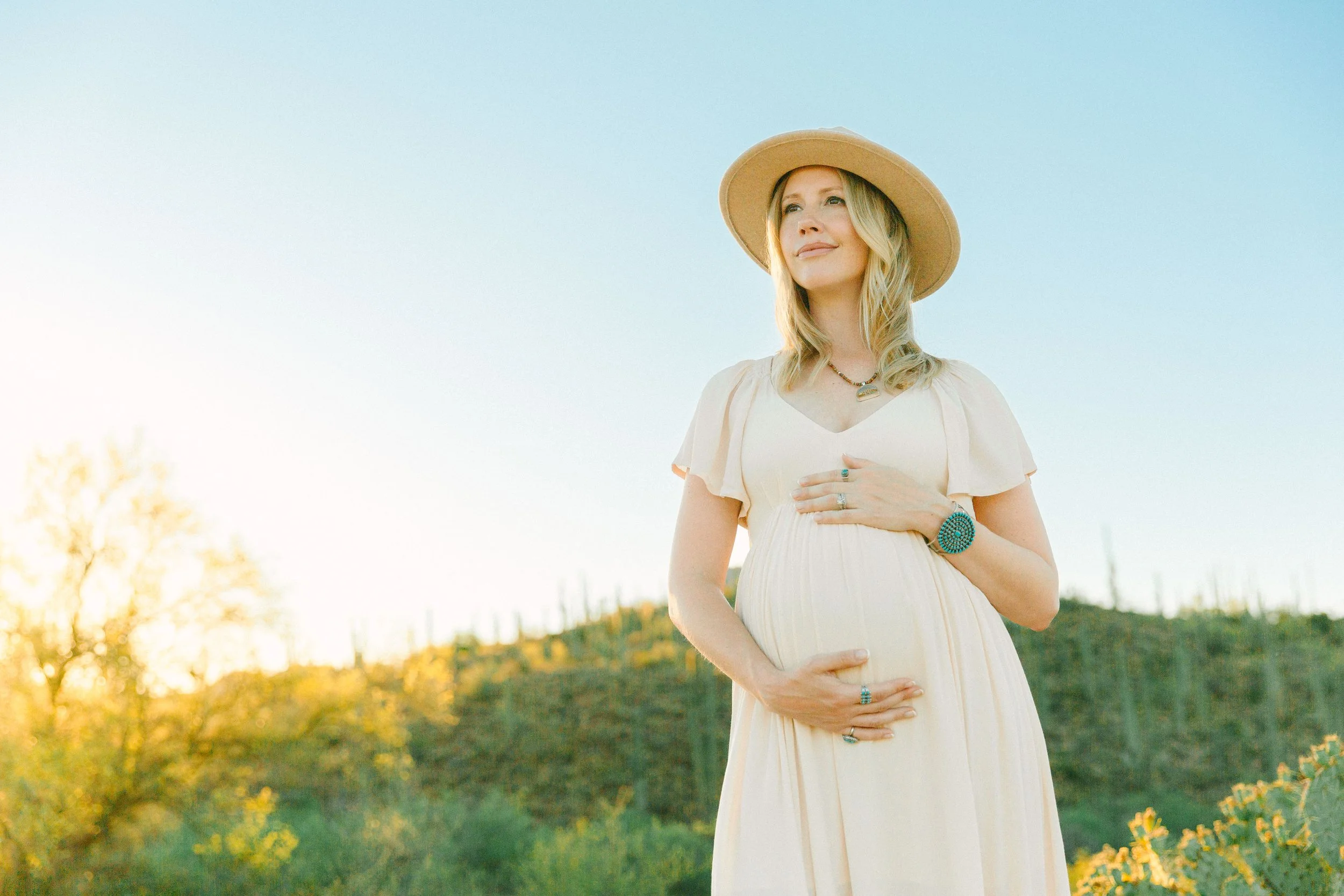 A pregnant woman outdoors during sunset, wearing a wide-brimmed hat, a light-colored dress, jewelry, and smiling softly while holding her belly.