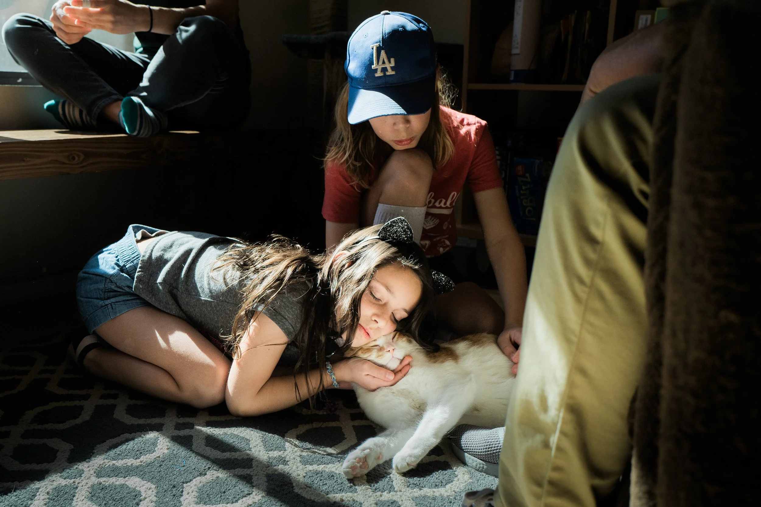 Children gathered around a large cat on a patterned rug, petting and cuddling it. One girl is lying down with her eyes closed, while another girl, wearing a blue LAPD baseball cap, is kneeling nearby, both showing affection for the cat.