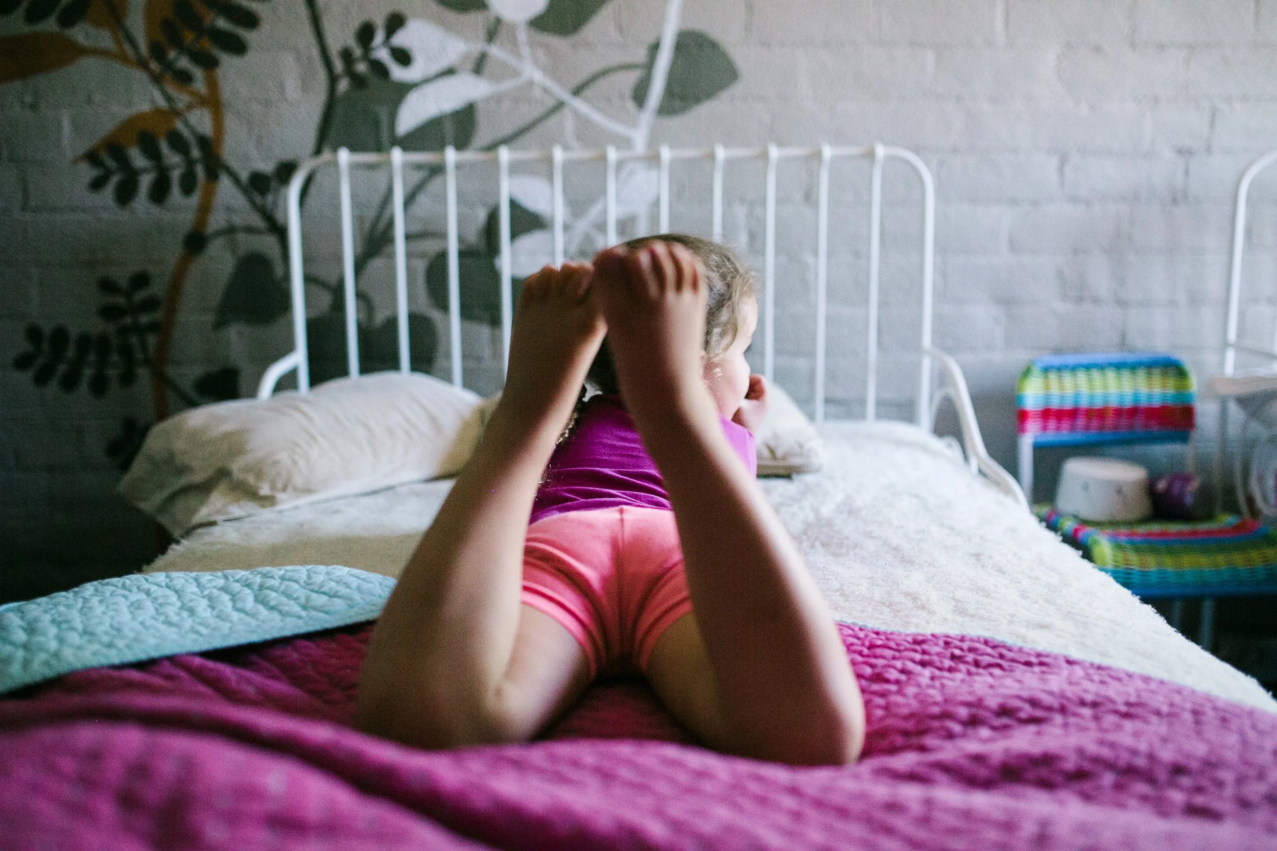 A young girl laying on her stomach on a bed, holding her head, with a colorful room background.